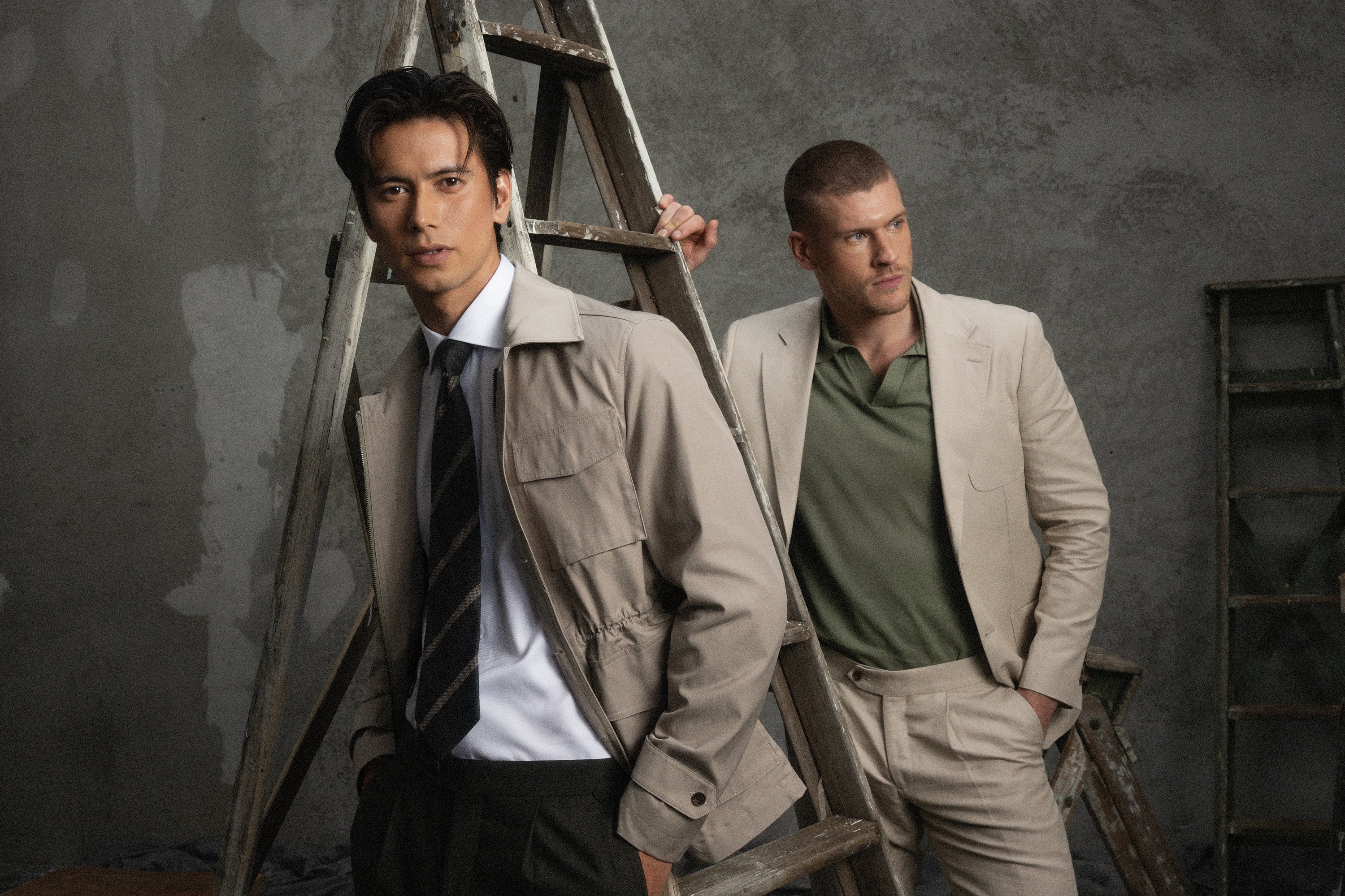 Two men in formal attire standing next to a ladder against a textured wall.