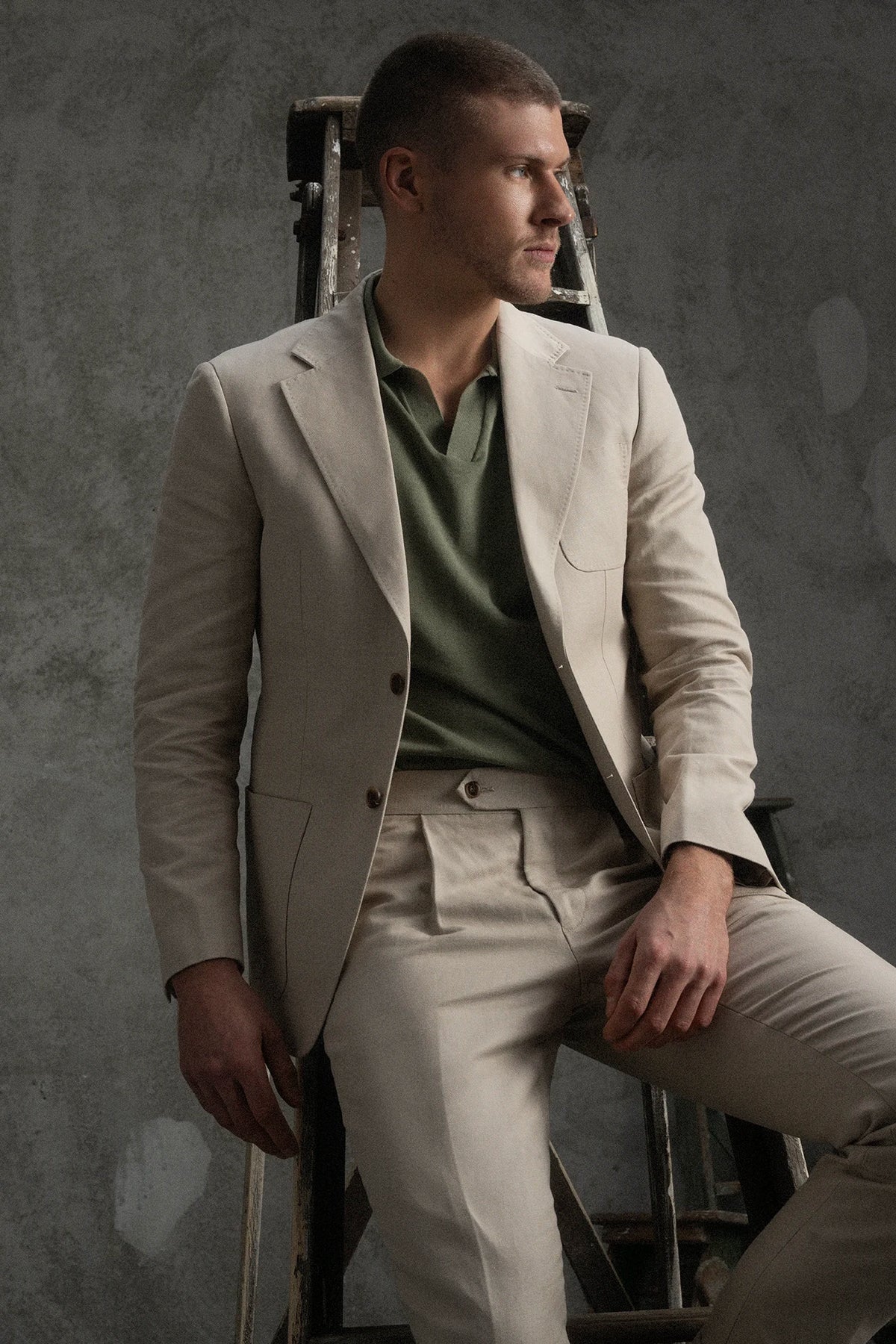 Man in a beige suit and green shirt sitting on a chair against a gray background
