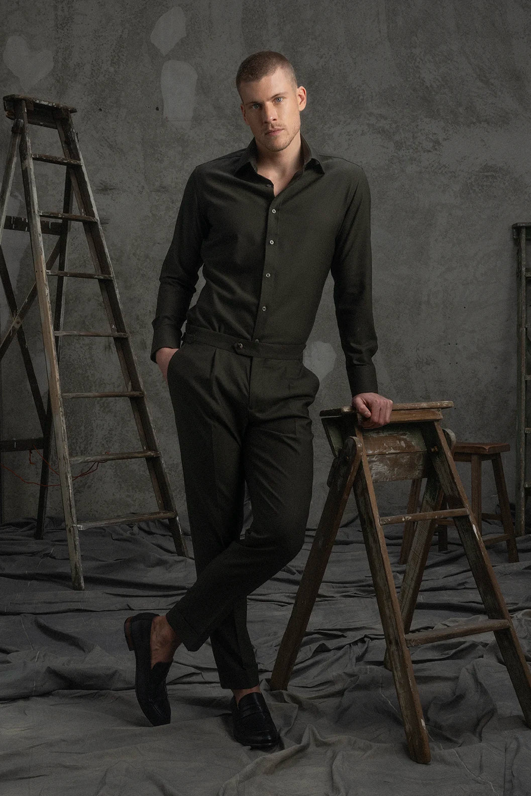 Man in a green shirt and pants standing in a studio with gray walls and a ladder.