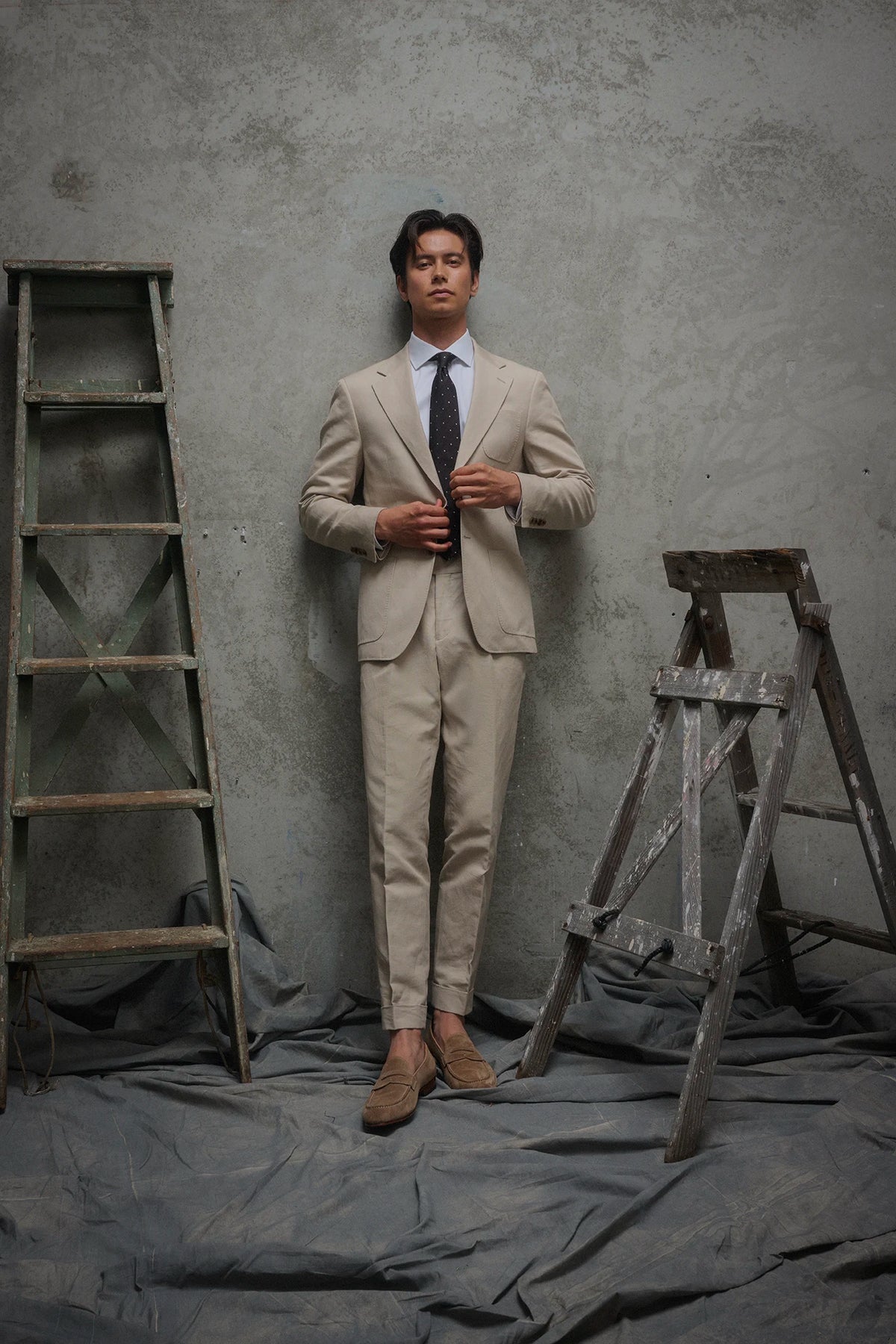 Man in a beige suit standing between two ladders against a gray textured wall.