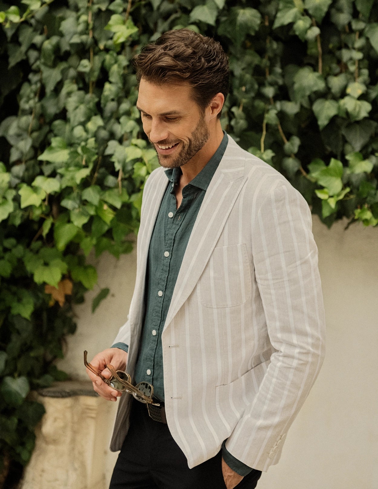 Man wearing a light-colored blazer over a green shirt, standing against a green leafy background.