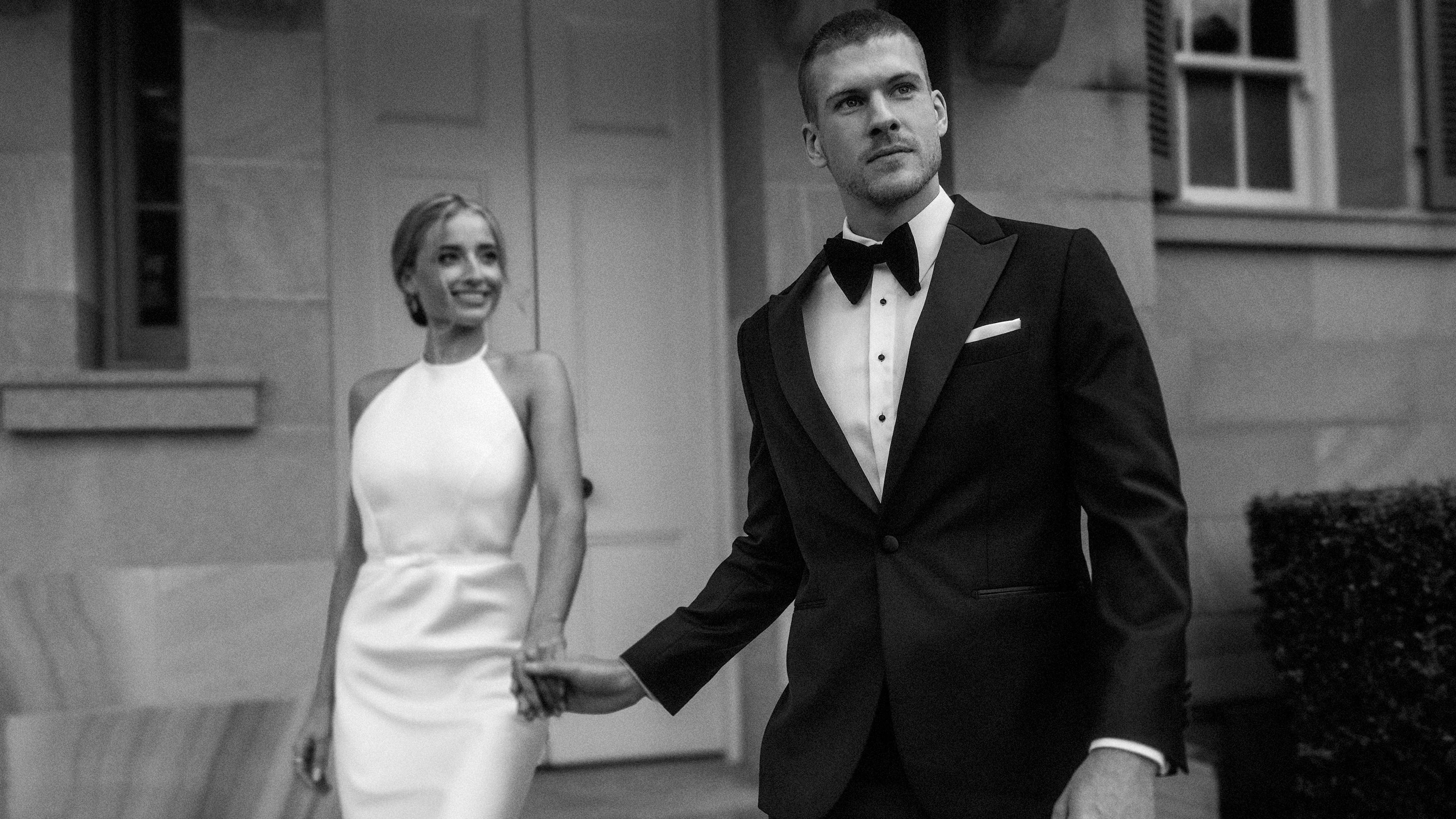 Man in a tuxedo and woman in a white dress standing together outdoors.