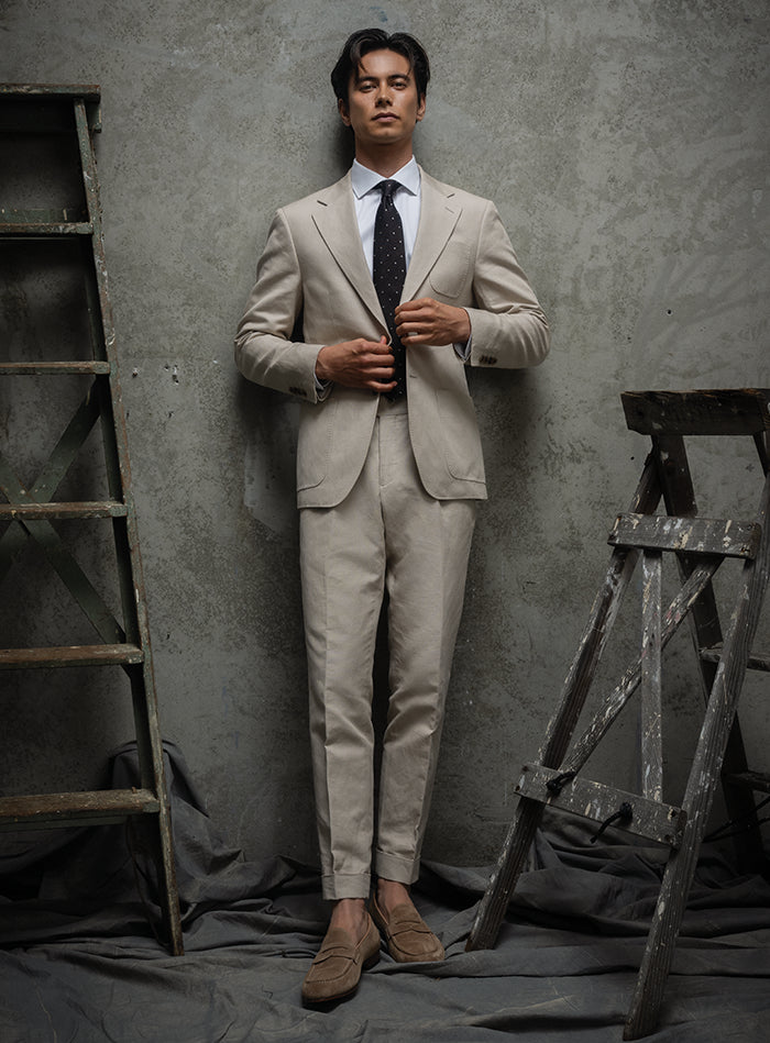 Man in a beige suit standing against a textured gray wall with wooden ladders.