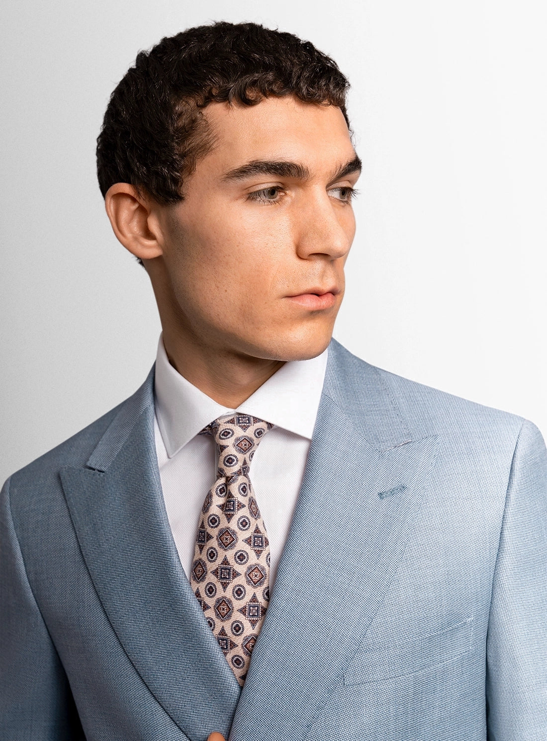 Man wearing a light blue suit with a patterned tie against a plain background