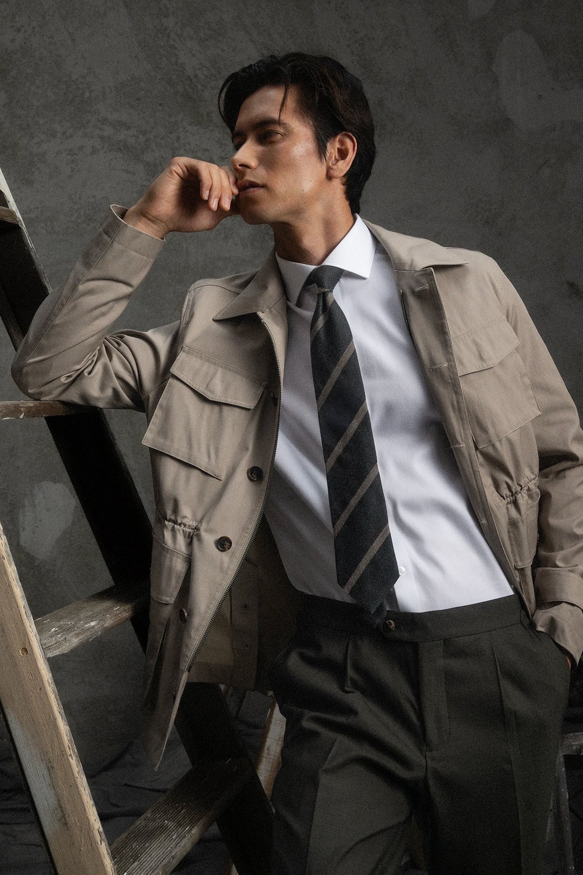 Man in a beige trench coat and striped tie leaning against a wall.