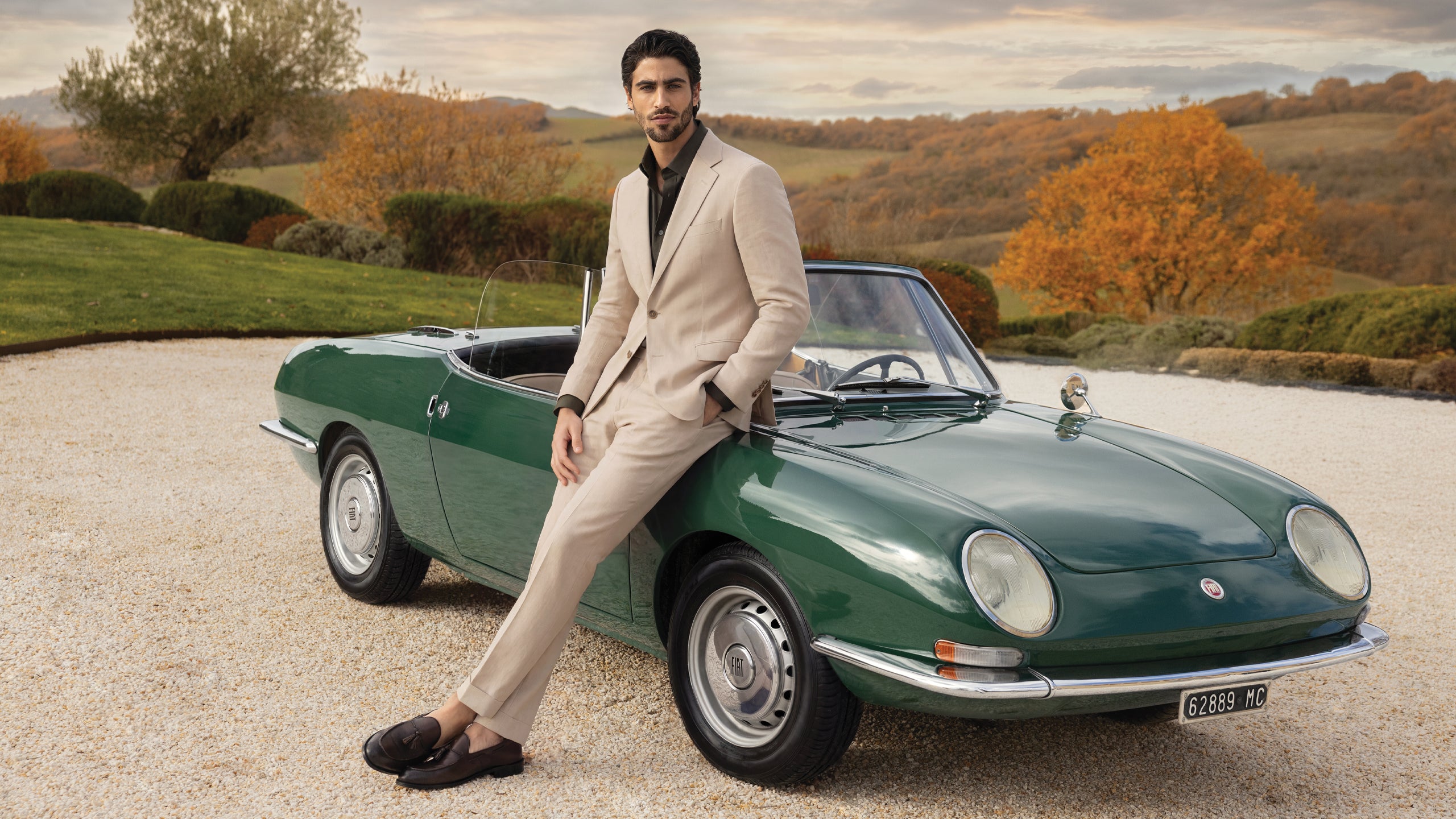 Man in a beige suit sitting on a vintage green car in a scenic outdoor setting