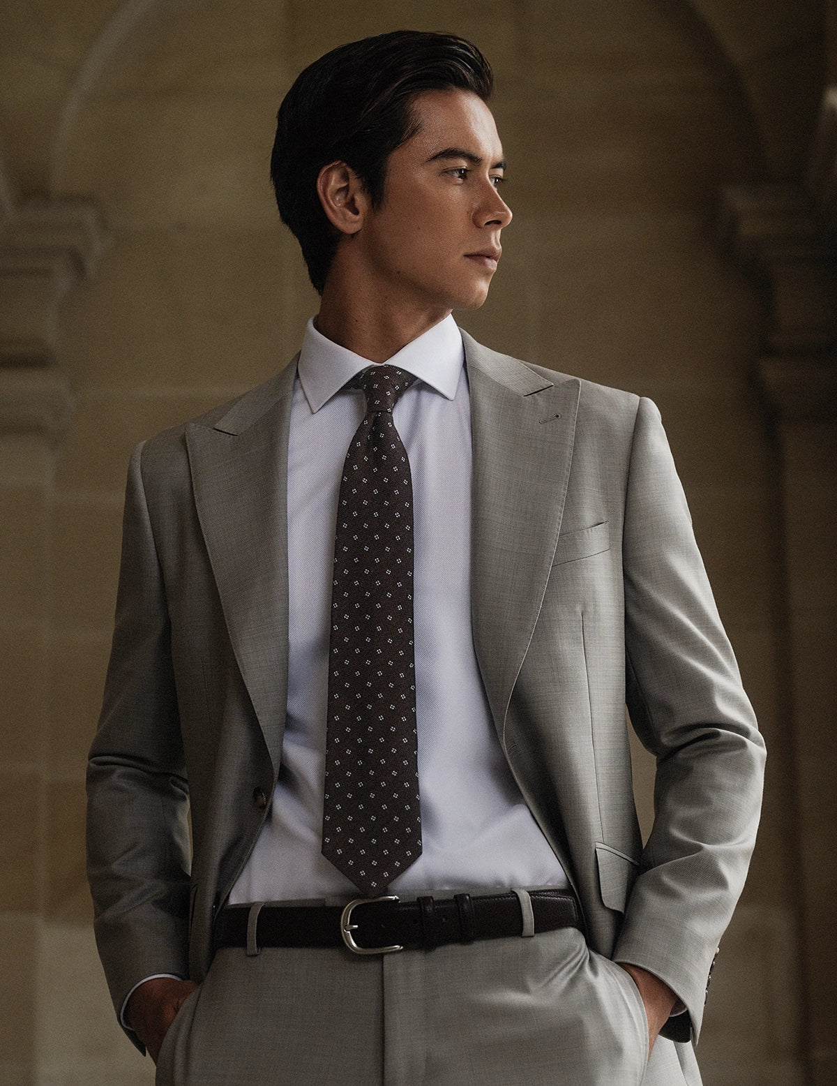 Man wearing a gray suit with a patterned tie against a stone wall background