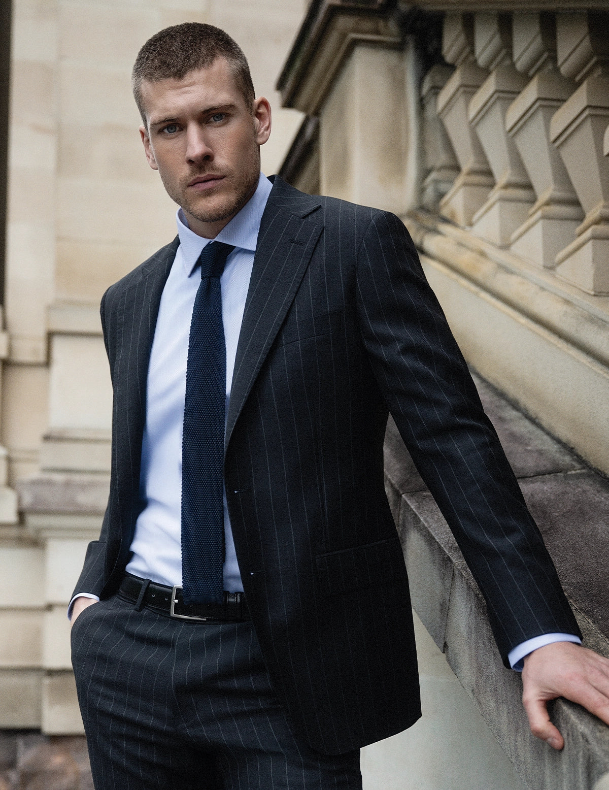Man in a dark pinstripe suit standing against a stone building.