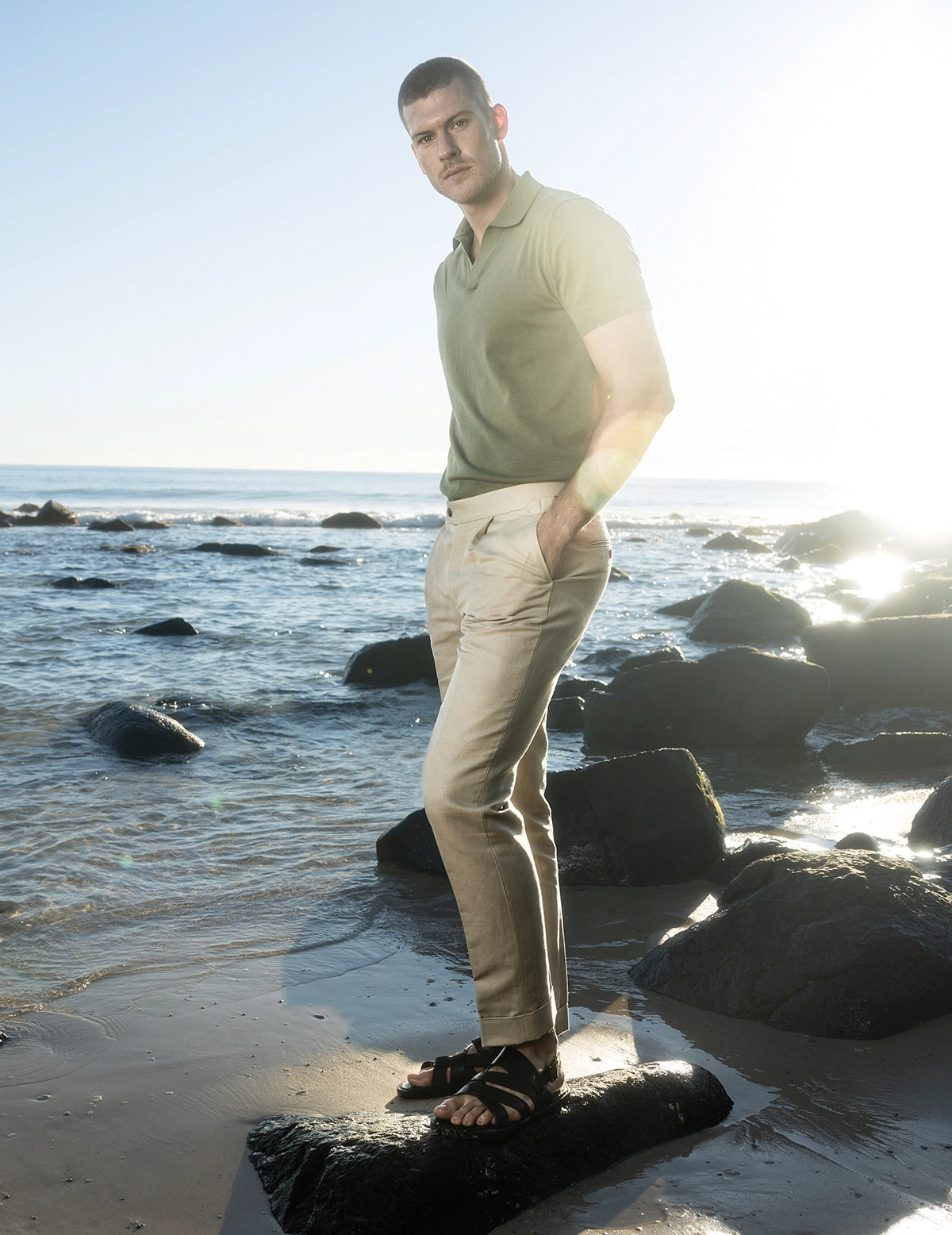 Man standing on a rocky beach wearing a light green polo shirt and beige pants.
