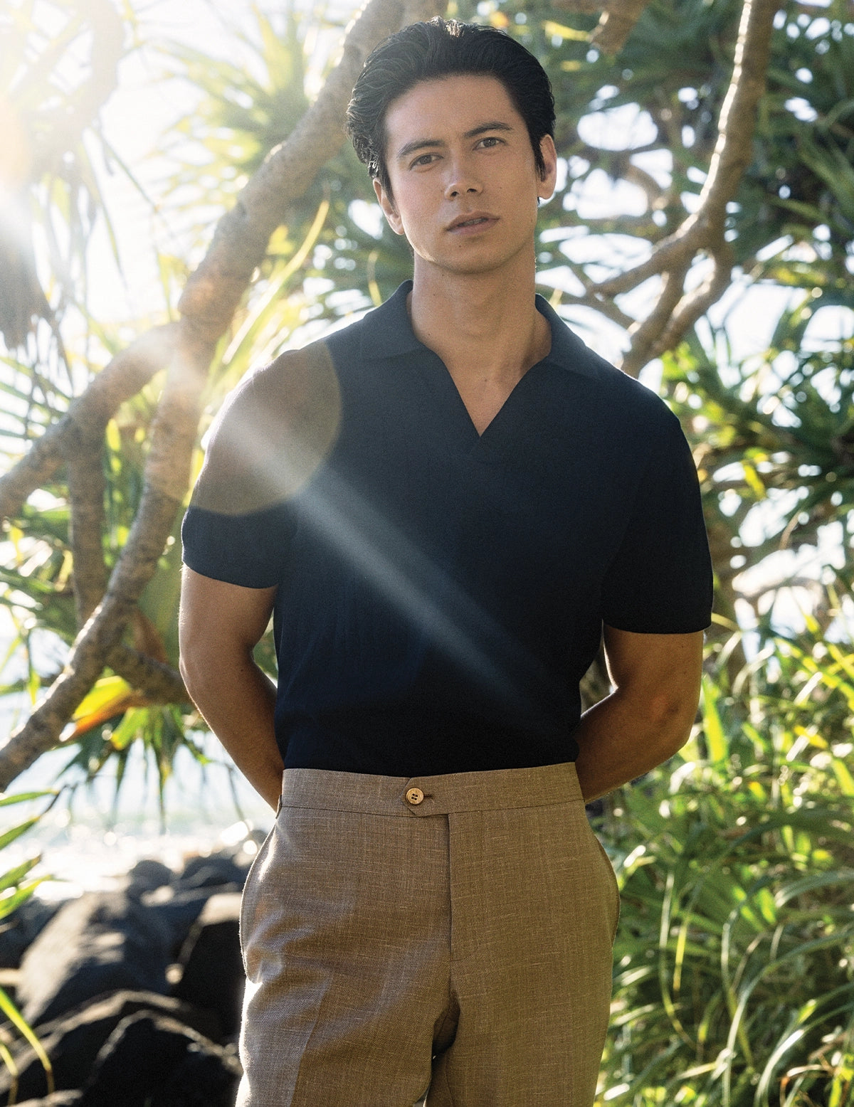 Man wearing a black shirt and beige pants standing in a natural setting with trees.