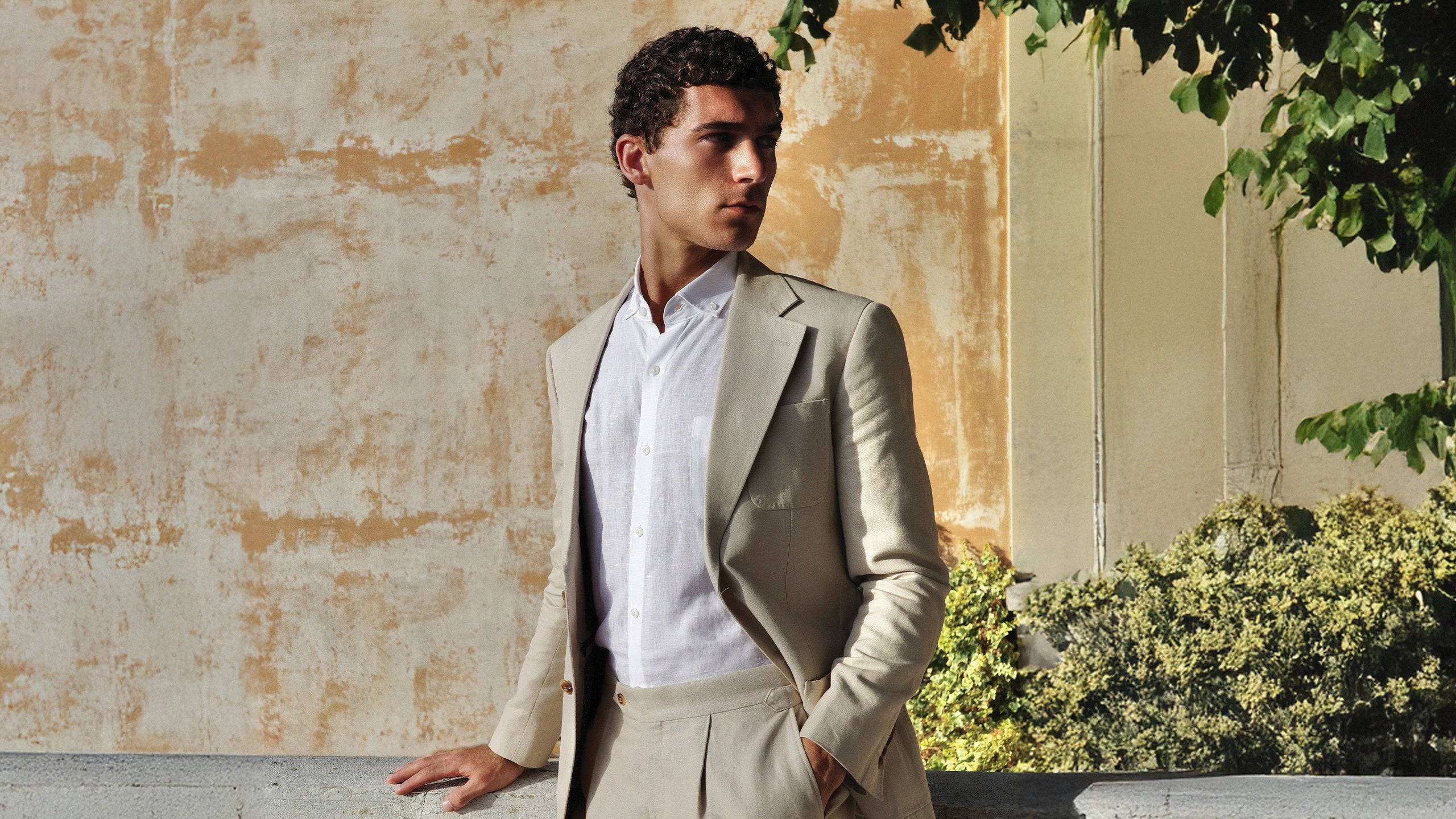 Man in a beige suit standing outdoors with a textured wall and greenery in the background