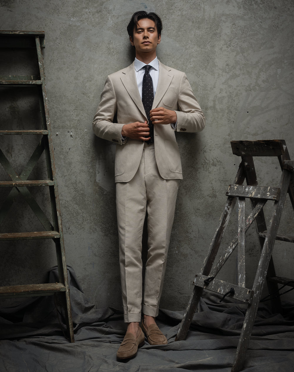 Man in a beige suit standing against a textured wall with wooden ladders.