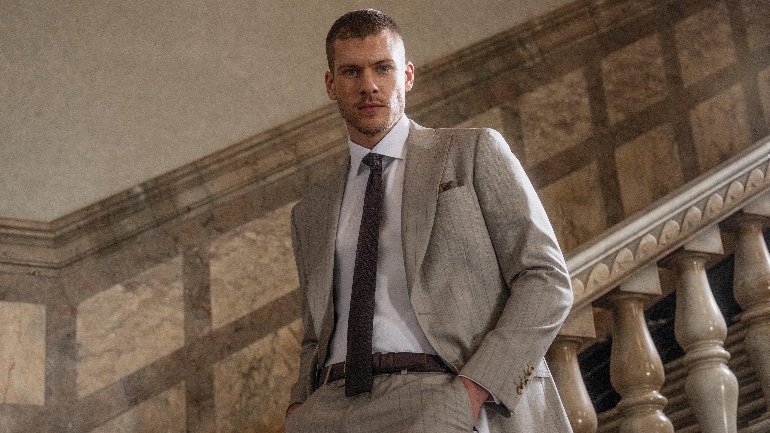 Man in a beige suit standing on a staircase with stone walls.