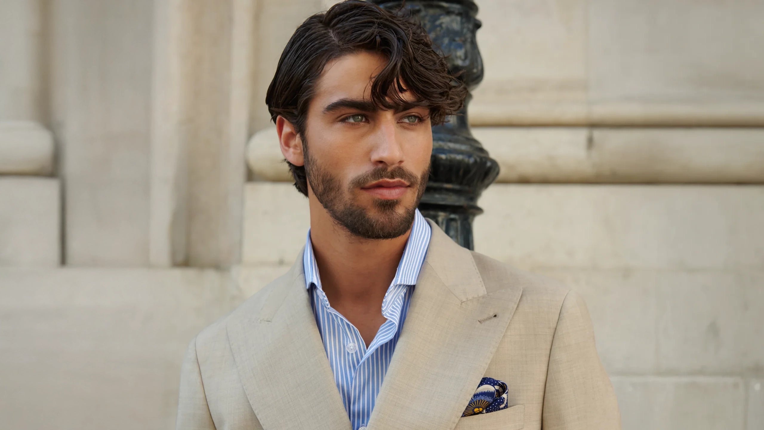 Man in a beige suit standing outdoors with classical architecture in the background