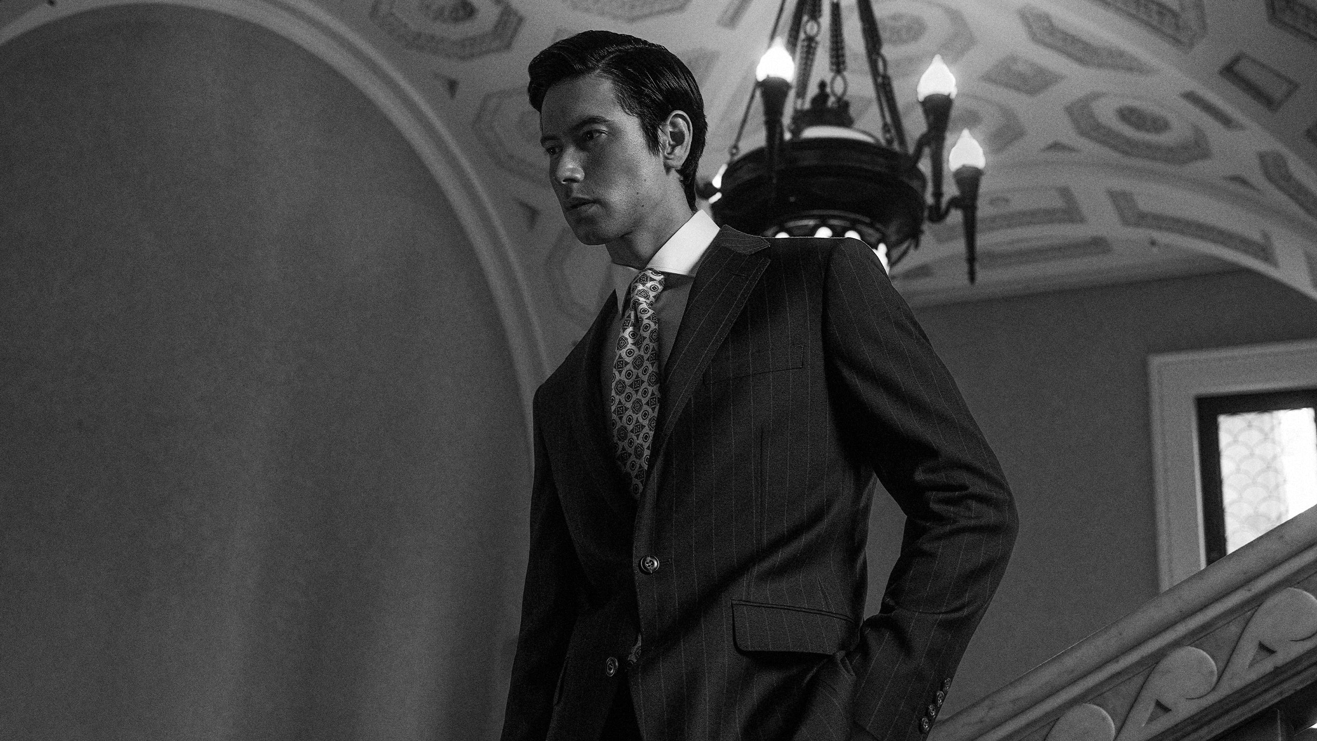 Man in a suit standing in an elegant interior setting with a chandelier.