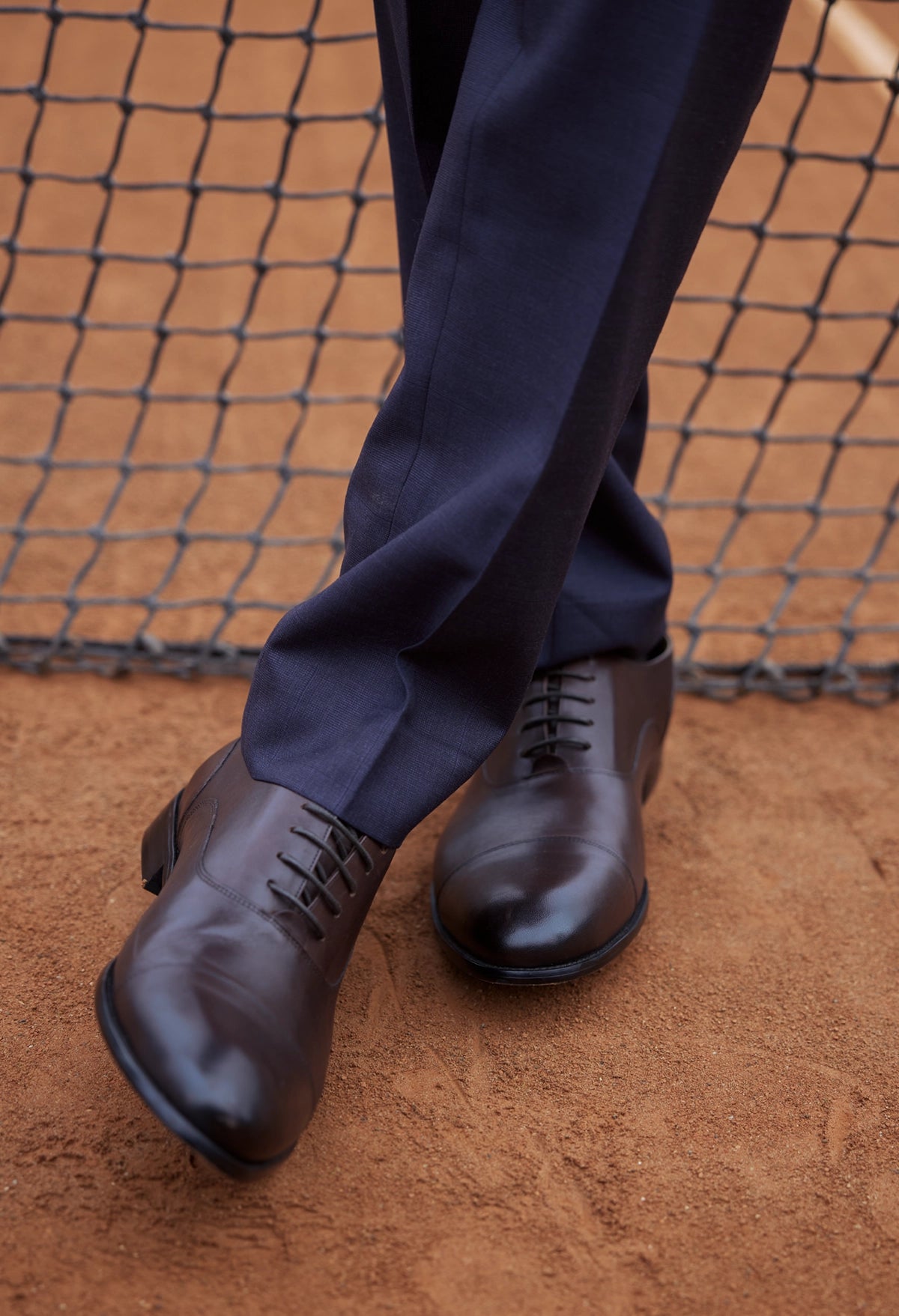 Dark blue dress pants and brown leather shoes on a dirt surface with a net in the background.