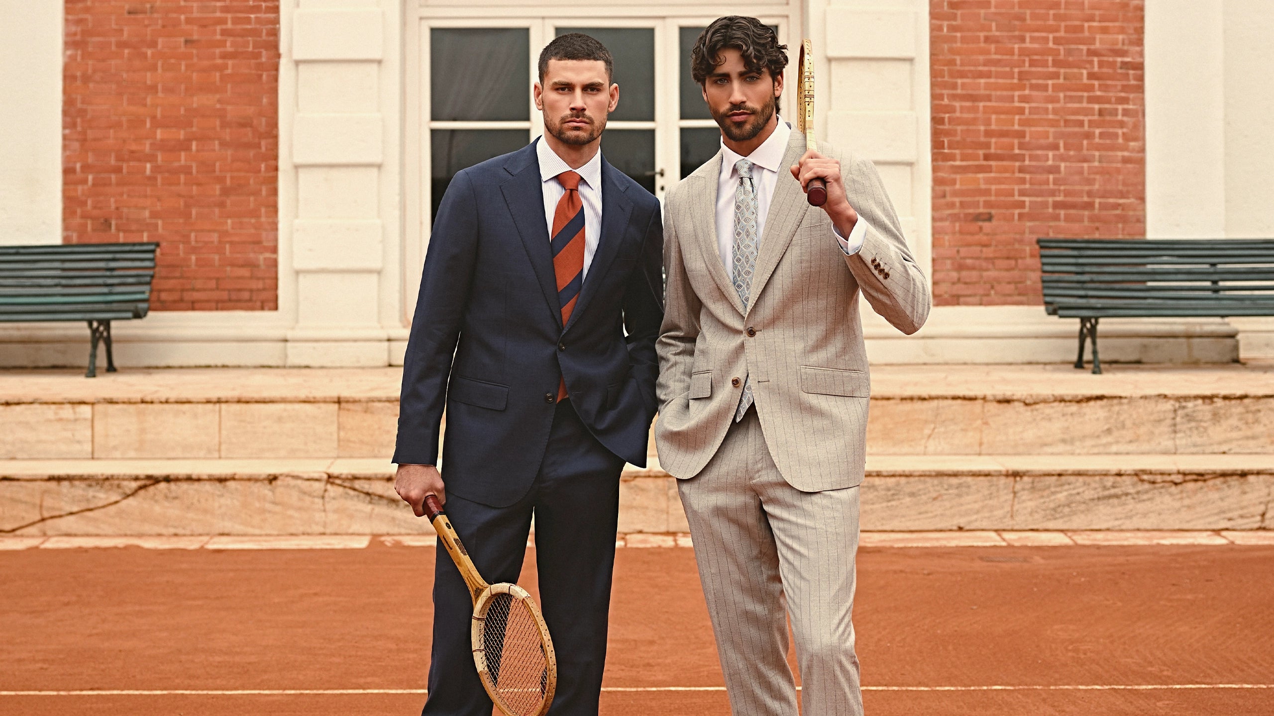 Two men in suits holding tennis rackets on a tennis court.