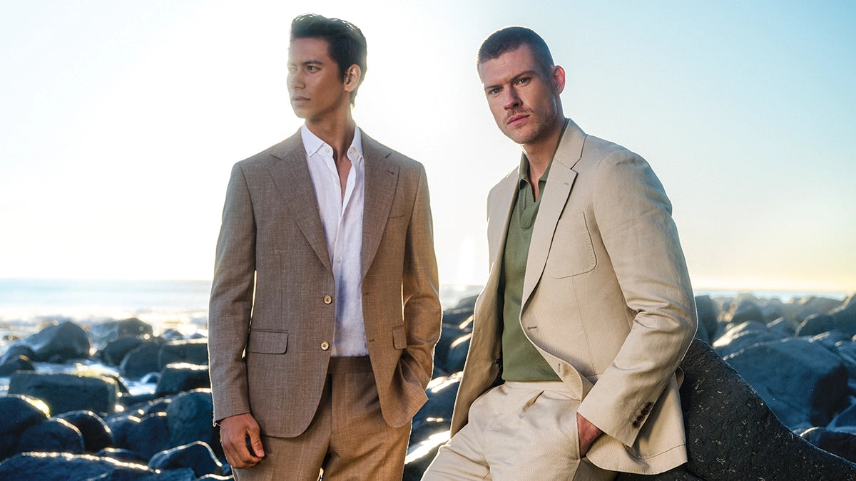 Two men in suits standing on a rocky beach with a clear sky.