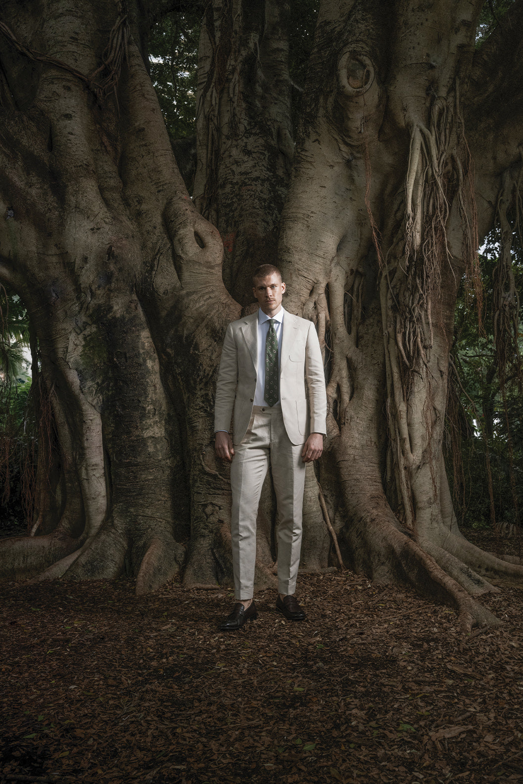 Man in a beige suit standing in front of a large tree with intricate root system.