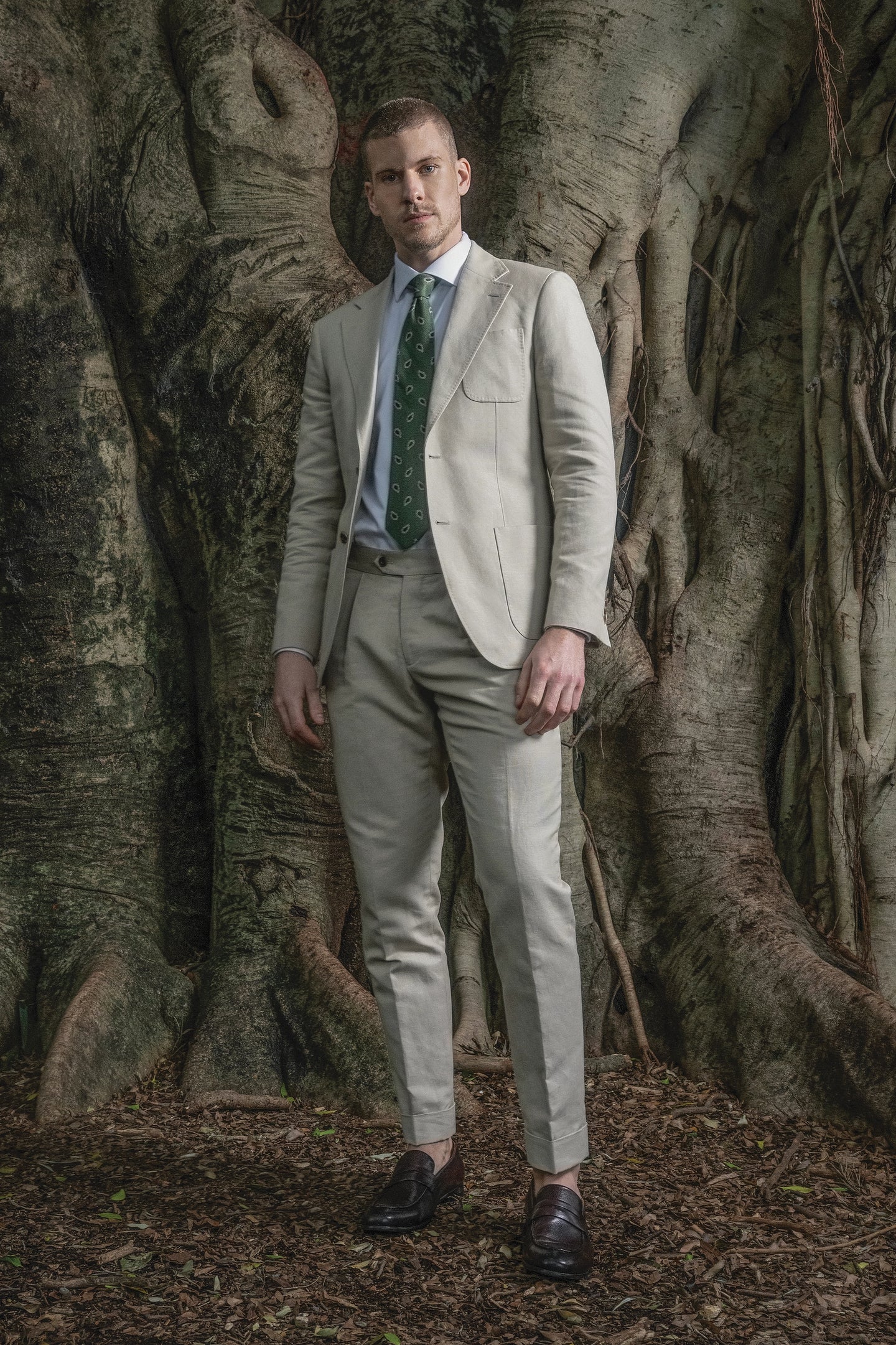 Man in a beige suit standing in front of large tree trunks
