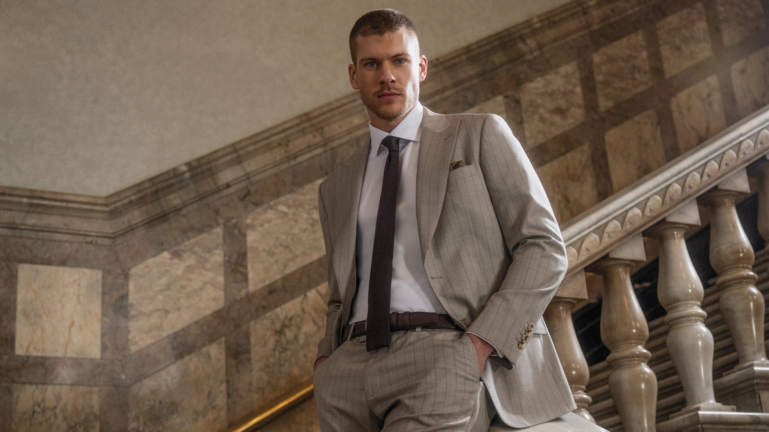 Man with beige suit and white suit standing behind stairs