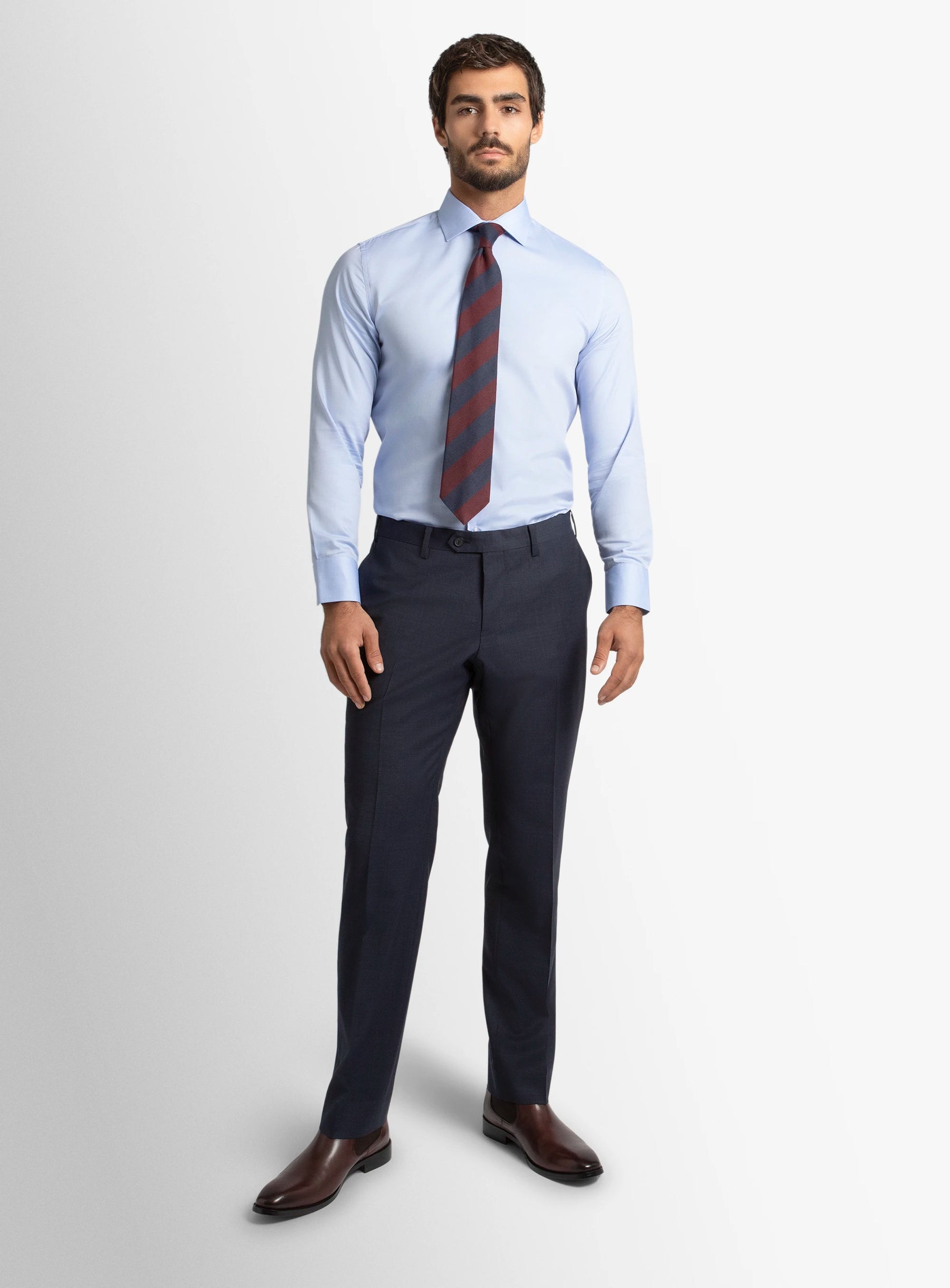 Man wearing a light blue dress shirt, dark tie, dark pants, and brown shoes on a white background