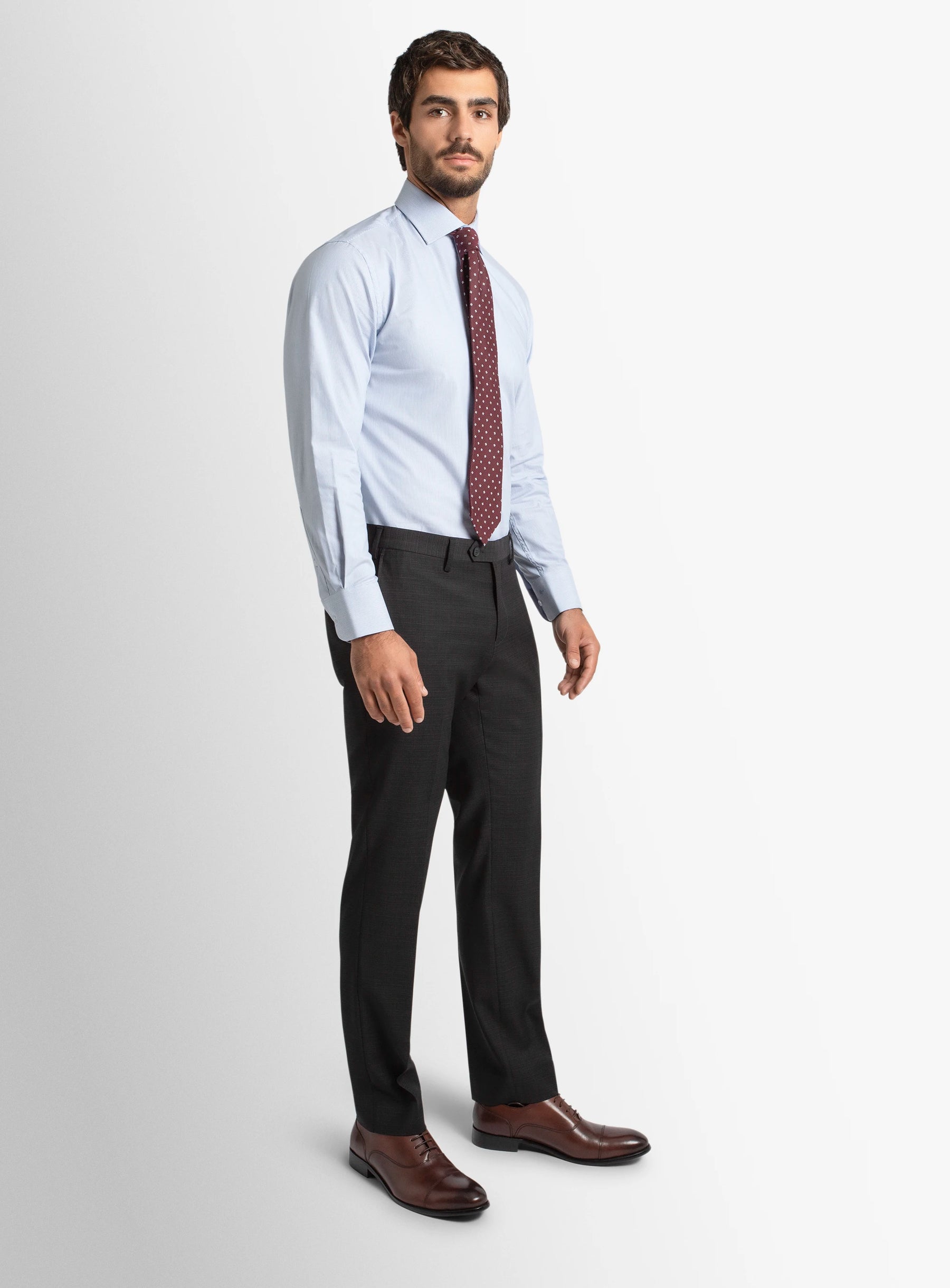 Man wearing a light blue dress shirt, dark tie, black pants, and brown shoes on a white background