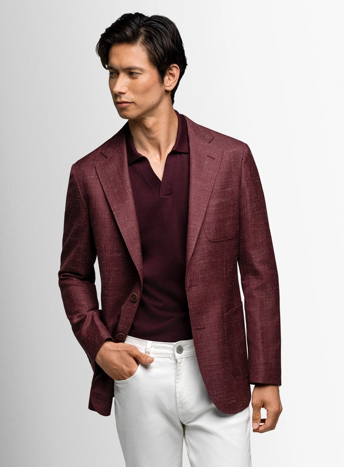 Man wearing a burgundy blazer over a dark shirt with white pants on a light gray background