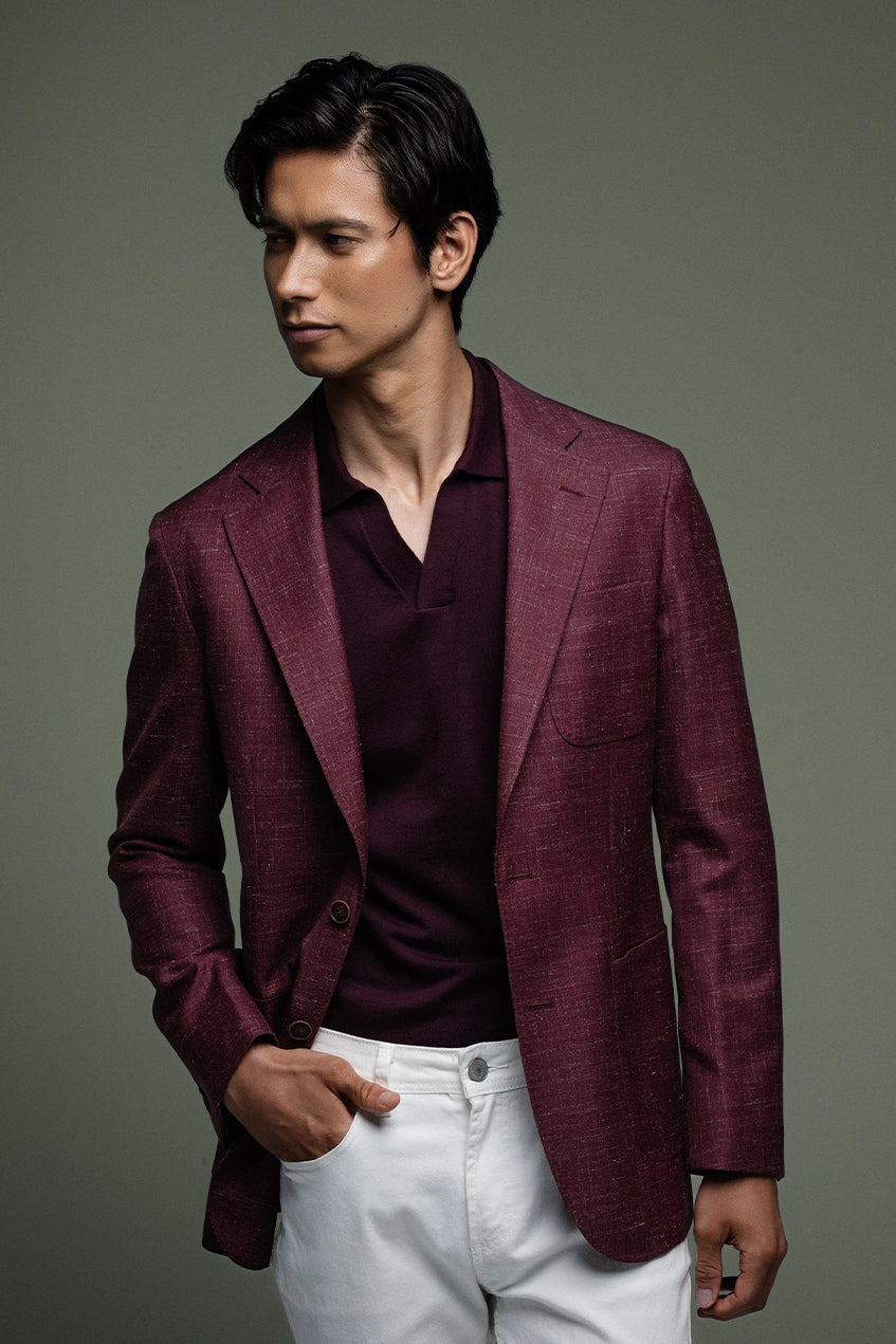 Man wearing a burgundy blazer over a dark shirt with white pants on a gray background