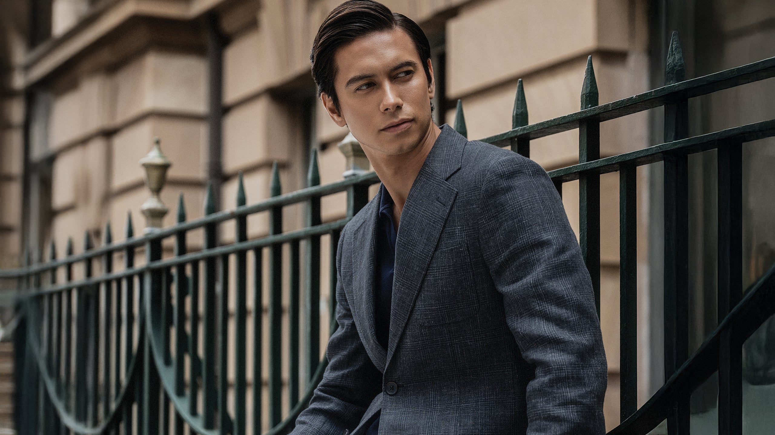 Man in a gray suit standing in front of a black metal gate with a building in the background