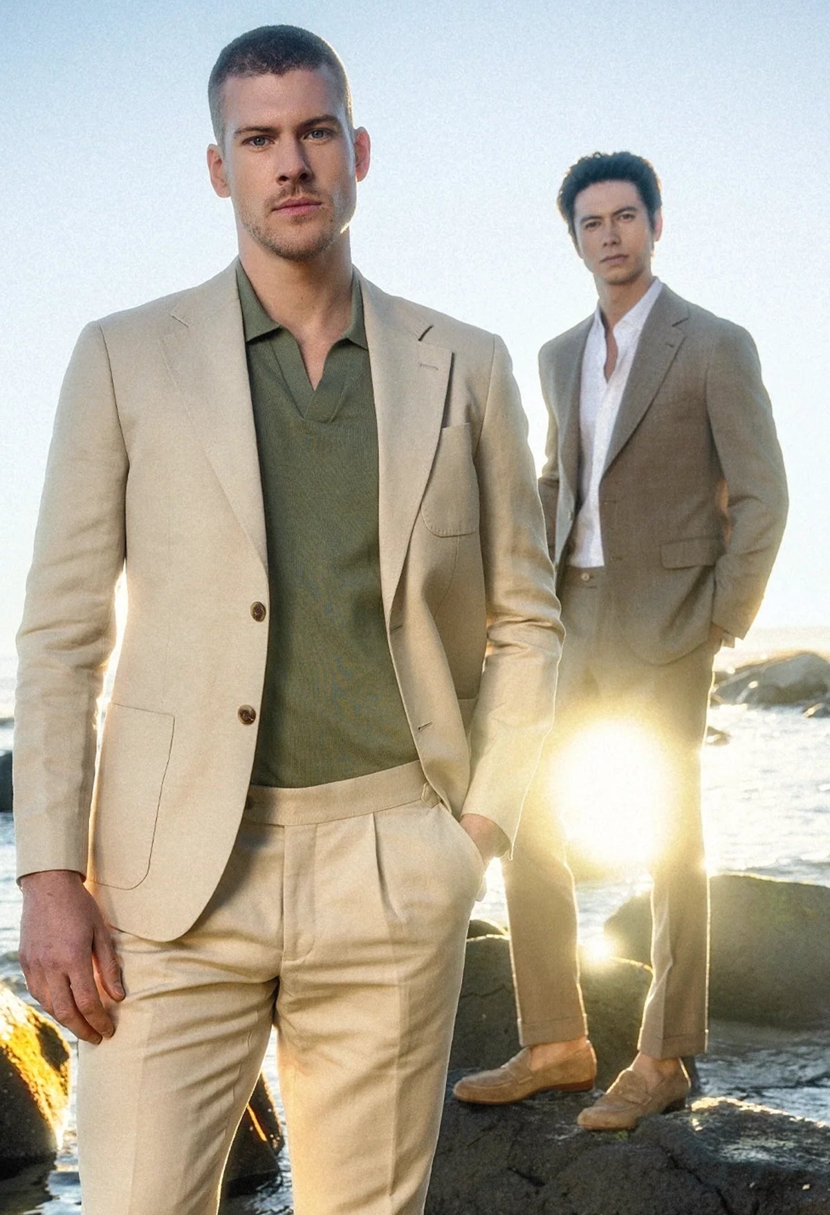 Two men in beige suits standing on a rocky beach with sunlight.