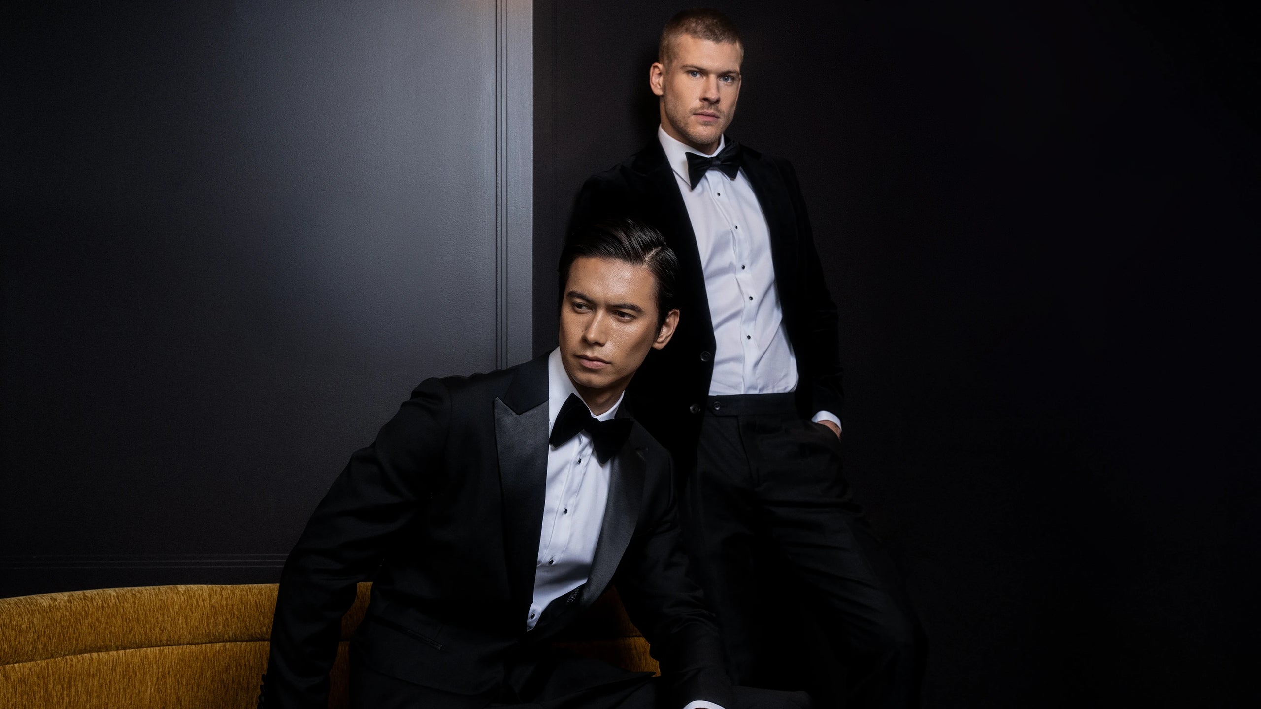 Two men in formal black suits sitting against a dark background