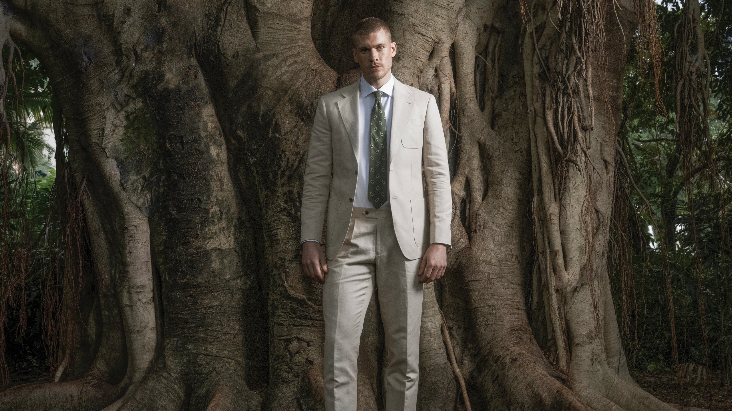 Man in a beige suit standing among large tree trunks