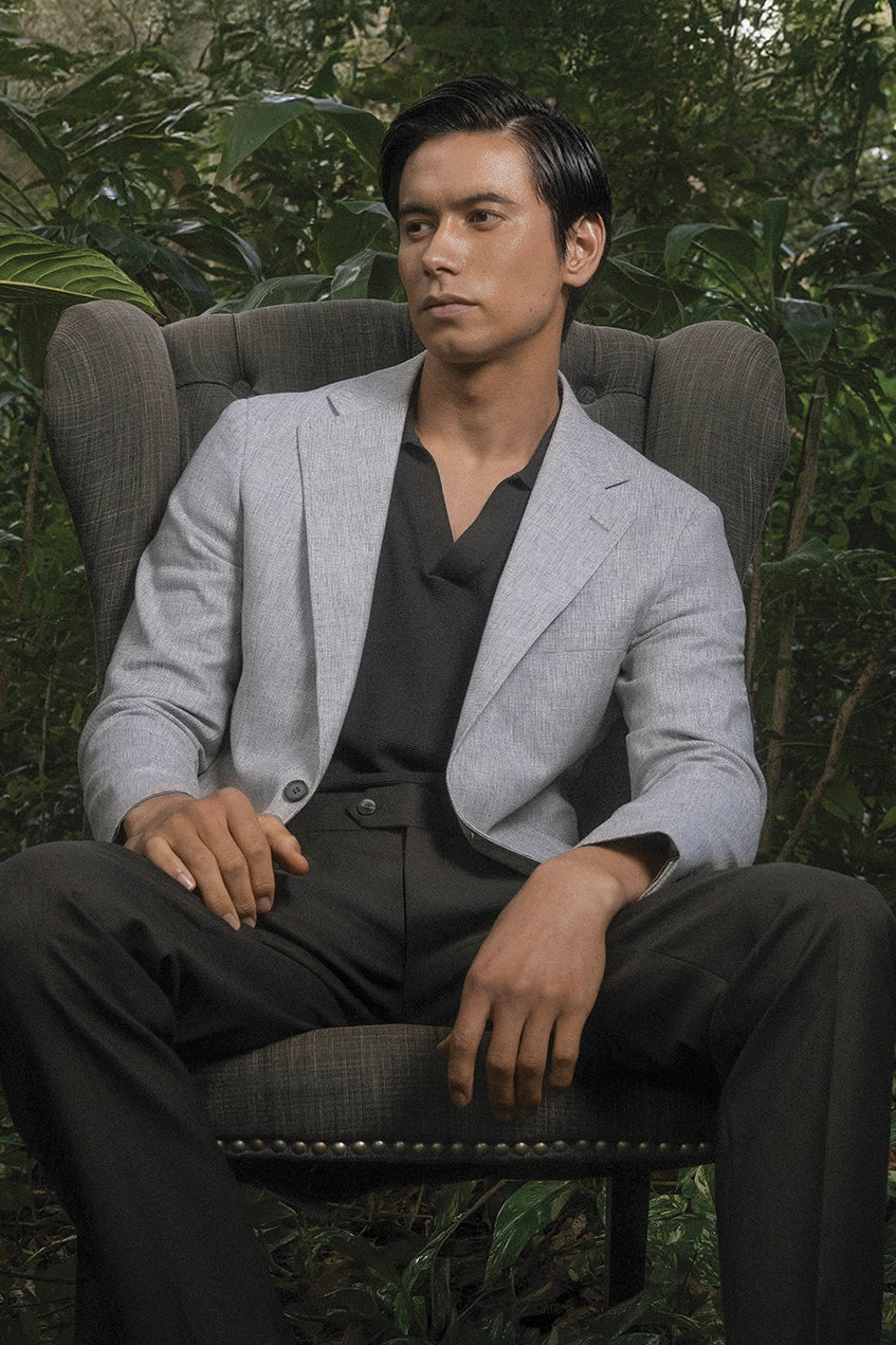 Man in a gray blazer sitting on a chair with greenery in the background
