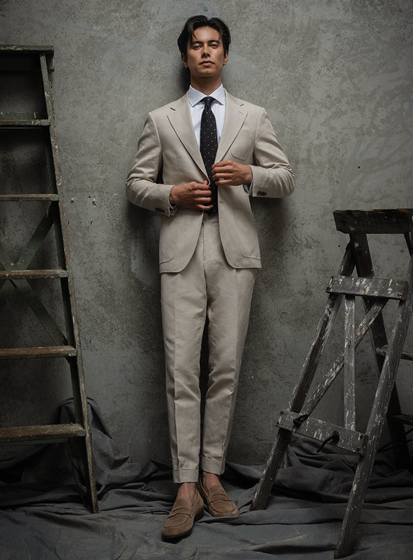 Man in a beige suit standing against a textured gray wall with wooden ladders.