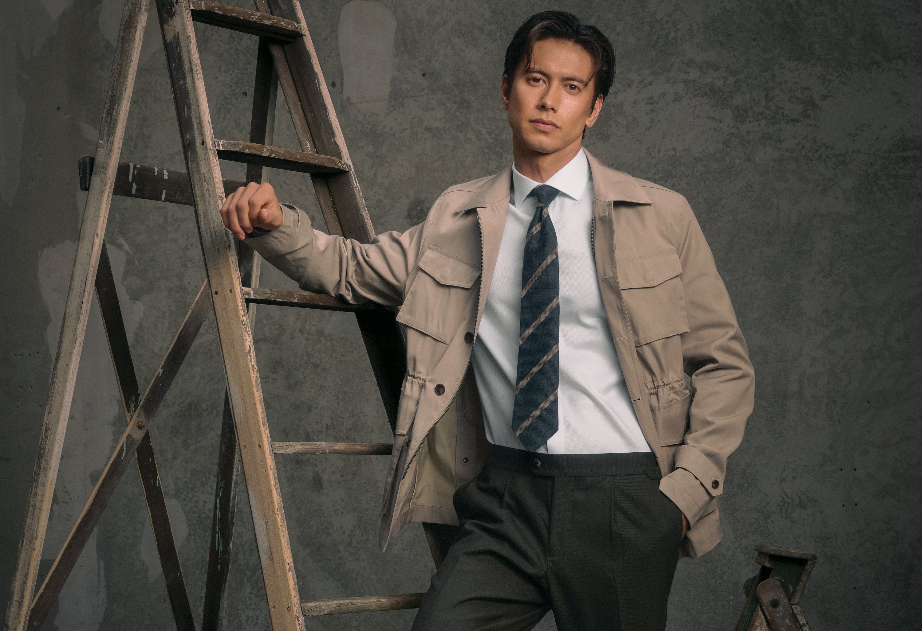 Man in a beige jacket and tie standing next to a wooden ladder against a gray background