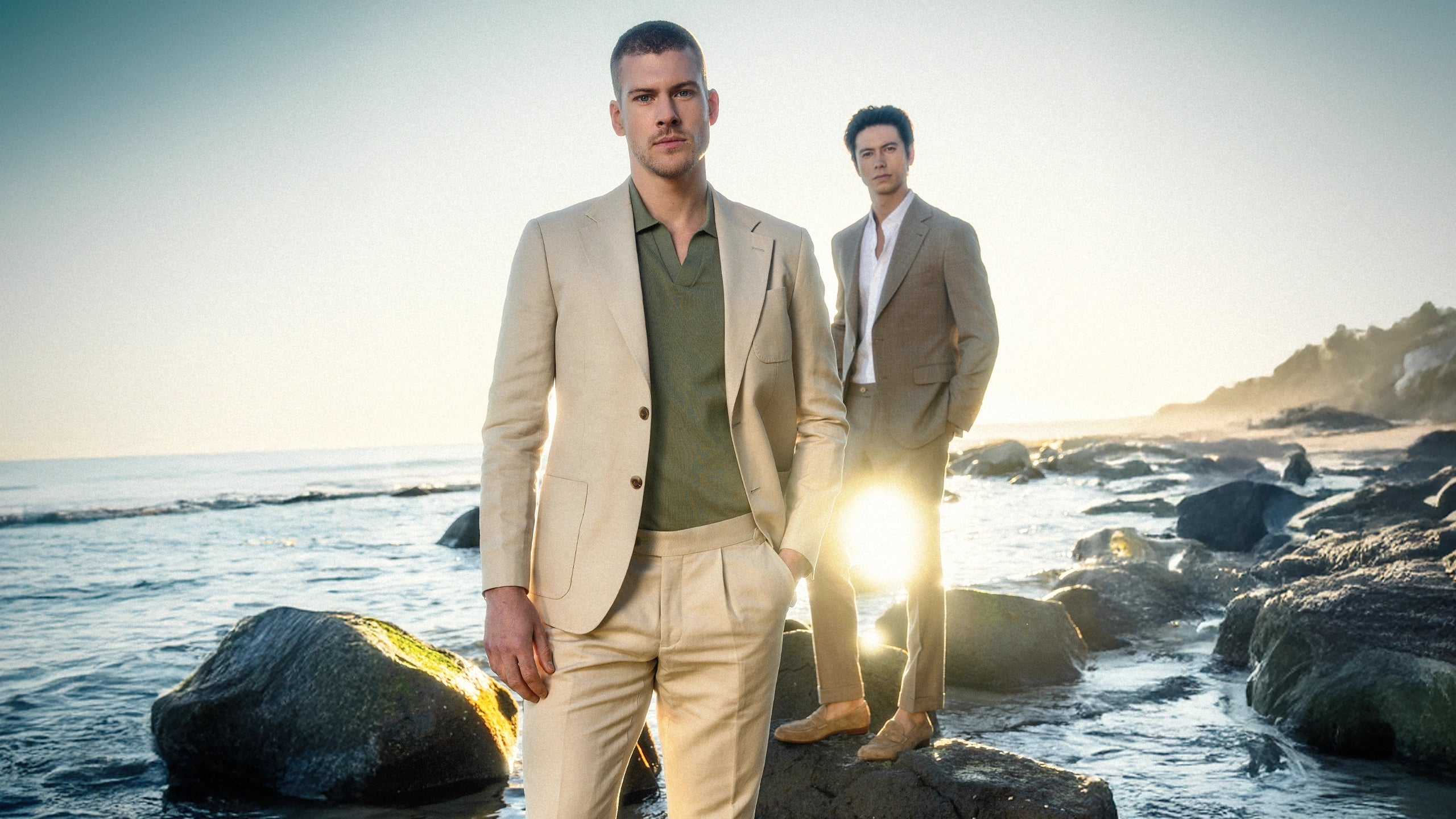 Two men in suits standing on a rocky beach with water and sky in the background.