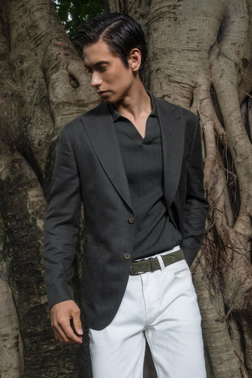 Man wearing a dark blazer, black shirt, and white pants standing against a tree.