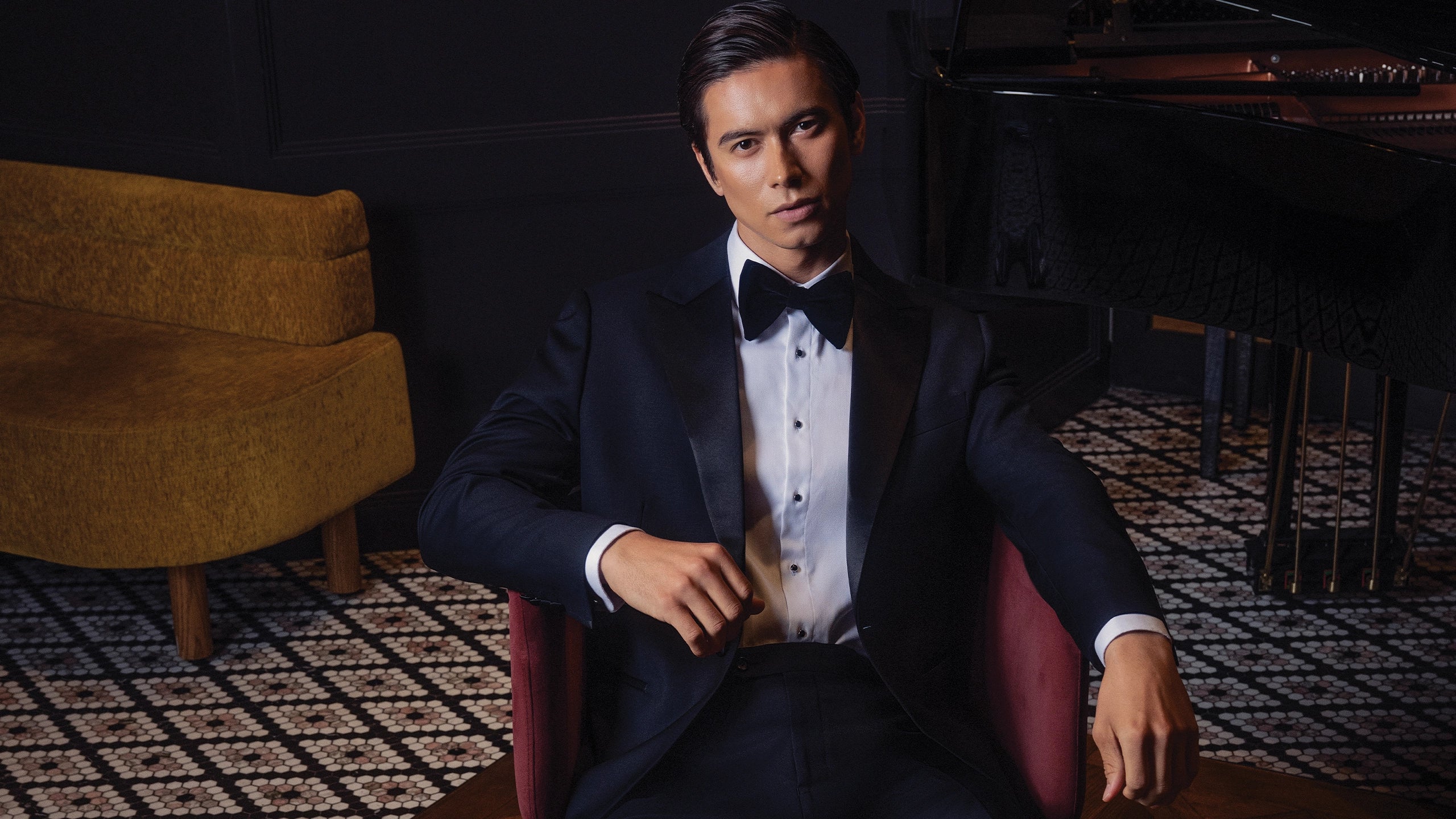 Man in a suit sitting in a room with a patterned rug and wooden furniture.