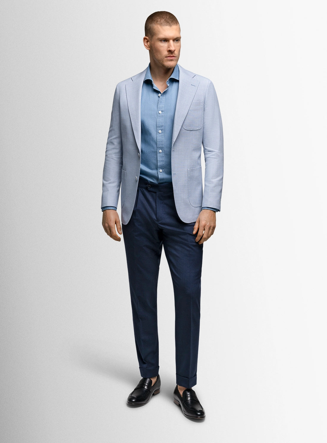 Man wearing a light blue blazer, light blue shirt, and dark blue pants on a white background