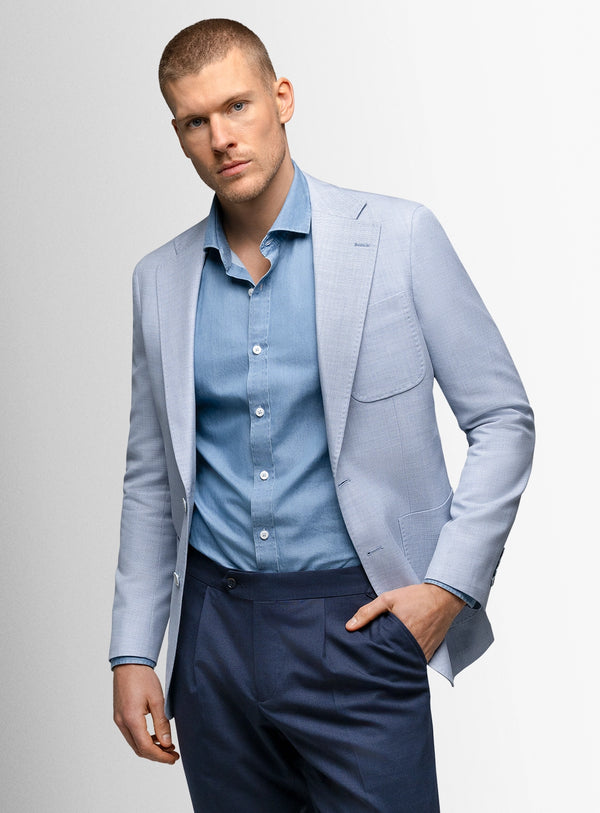 Man wearing a light blue blazer over a blue shirt with navy pants on a gray background