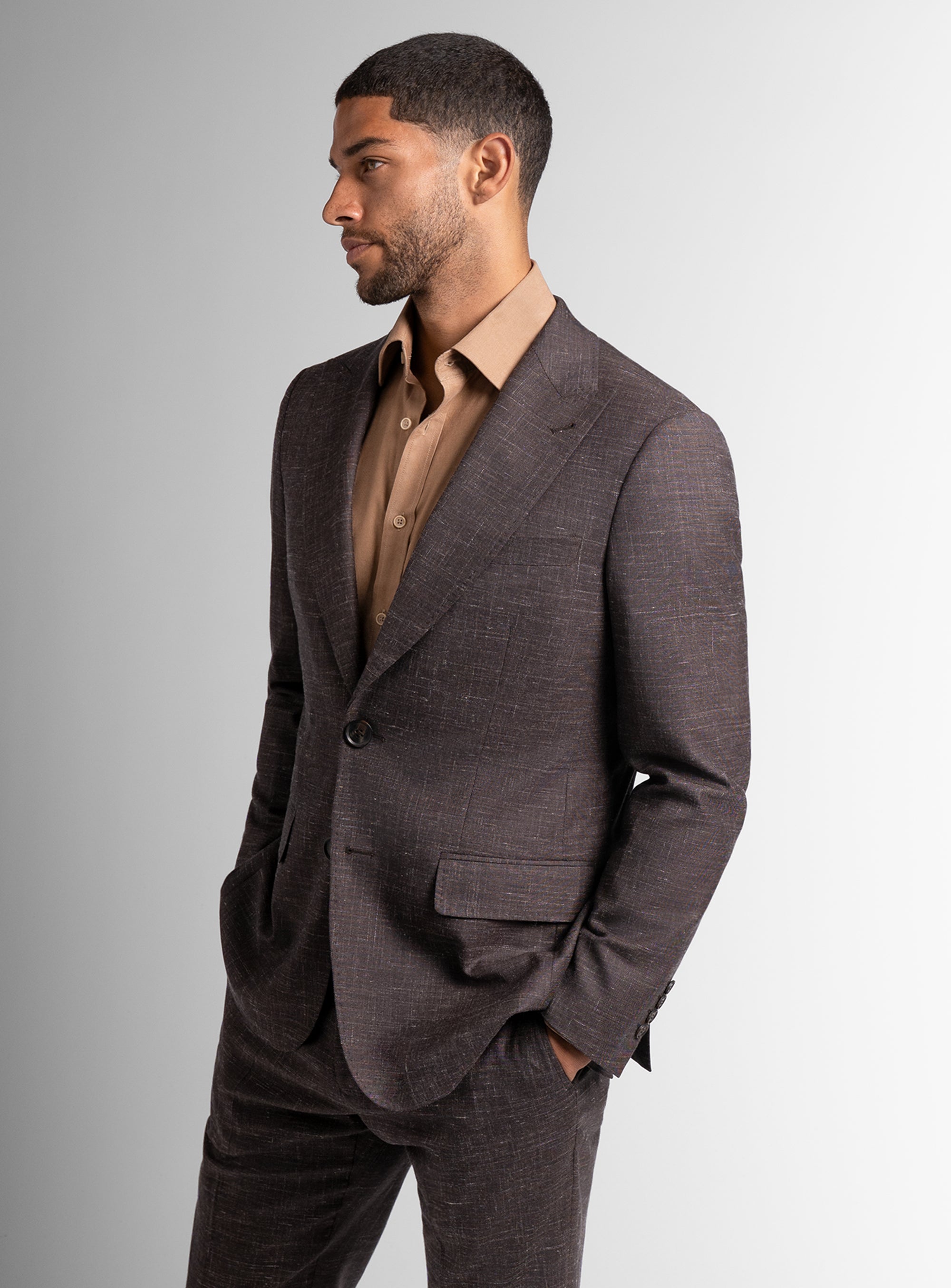 Man wearing a brown suit and beige shirt against a gray background