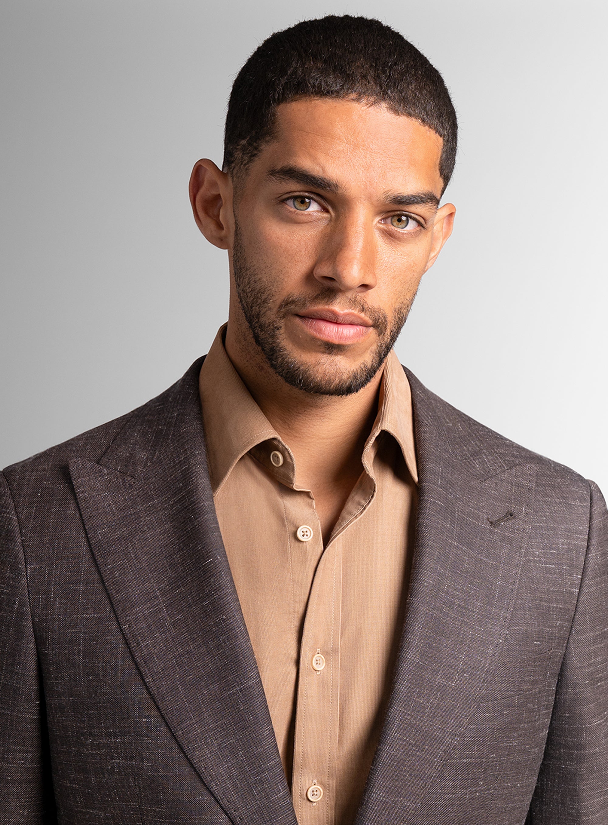 Man wearing a brown suit and beige shirt against a gray background
