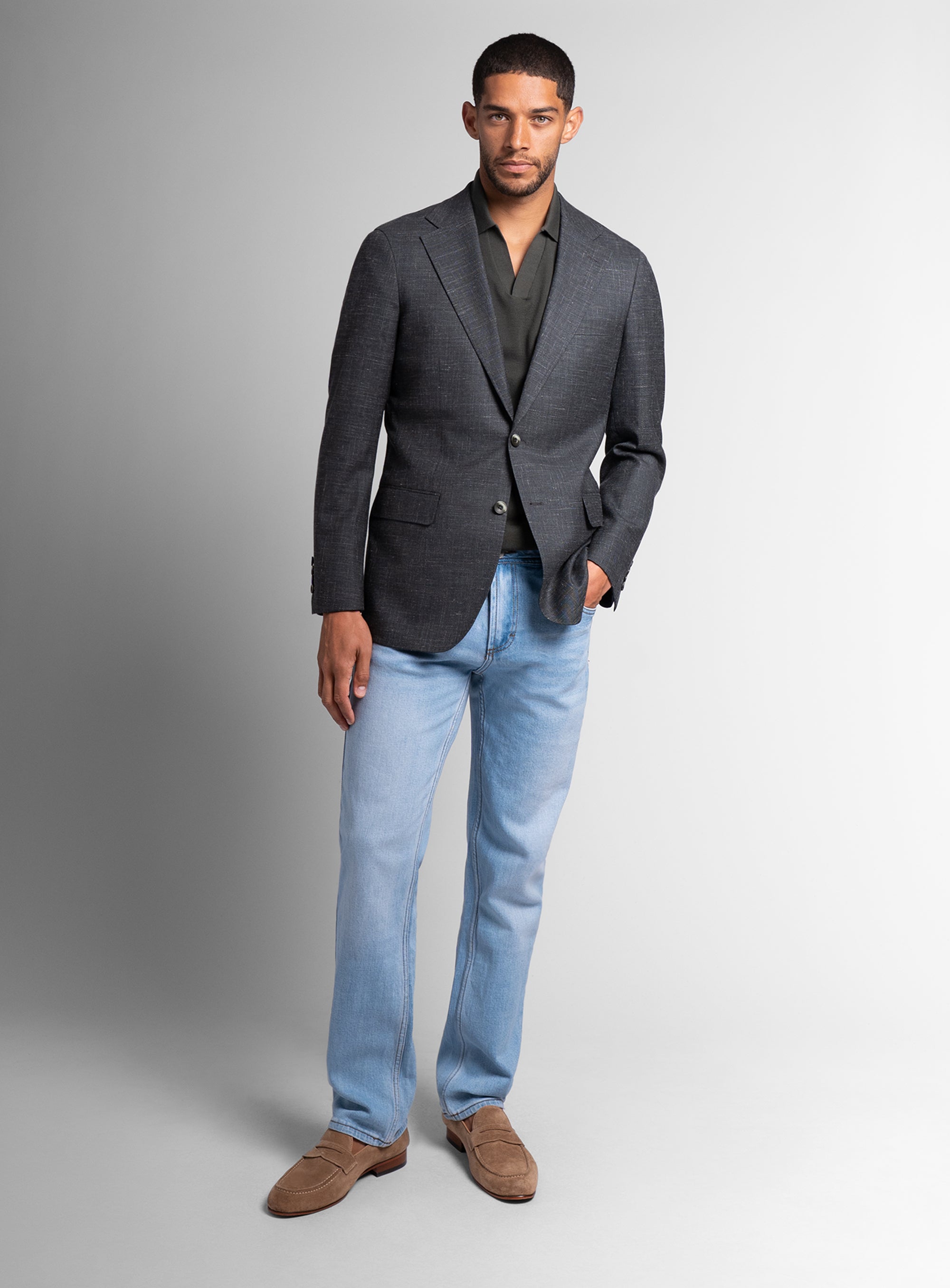 Man wearing a gray blazer, black shirt, light blue jeans, and brown shoes on a gray background