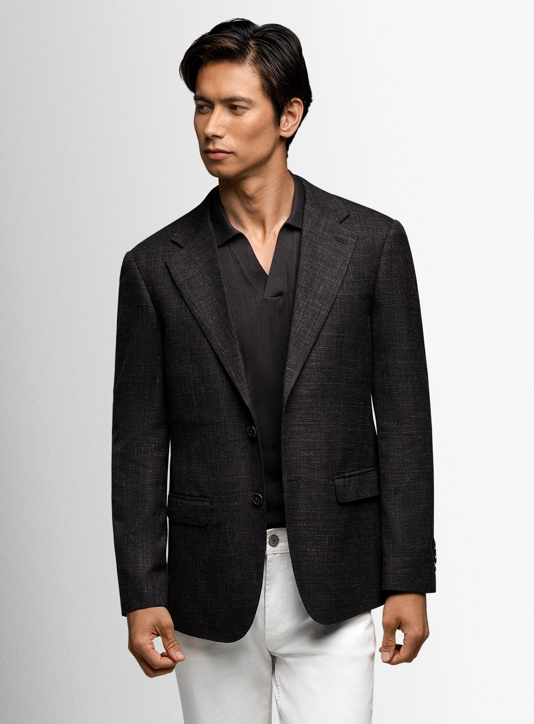 Man wearing a dark blazer over a black shirt with white pants on a light gray background