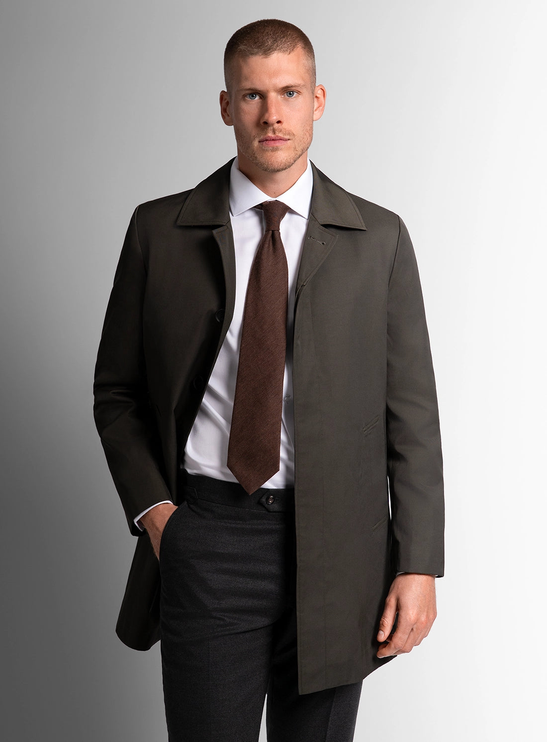Man wearing a dark coat over a suit with a brown tie on a gray background