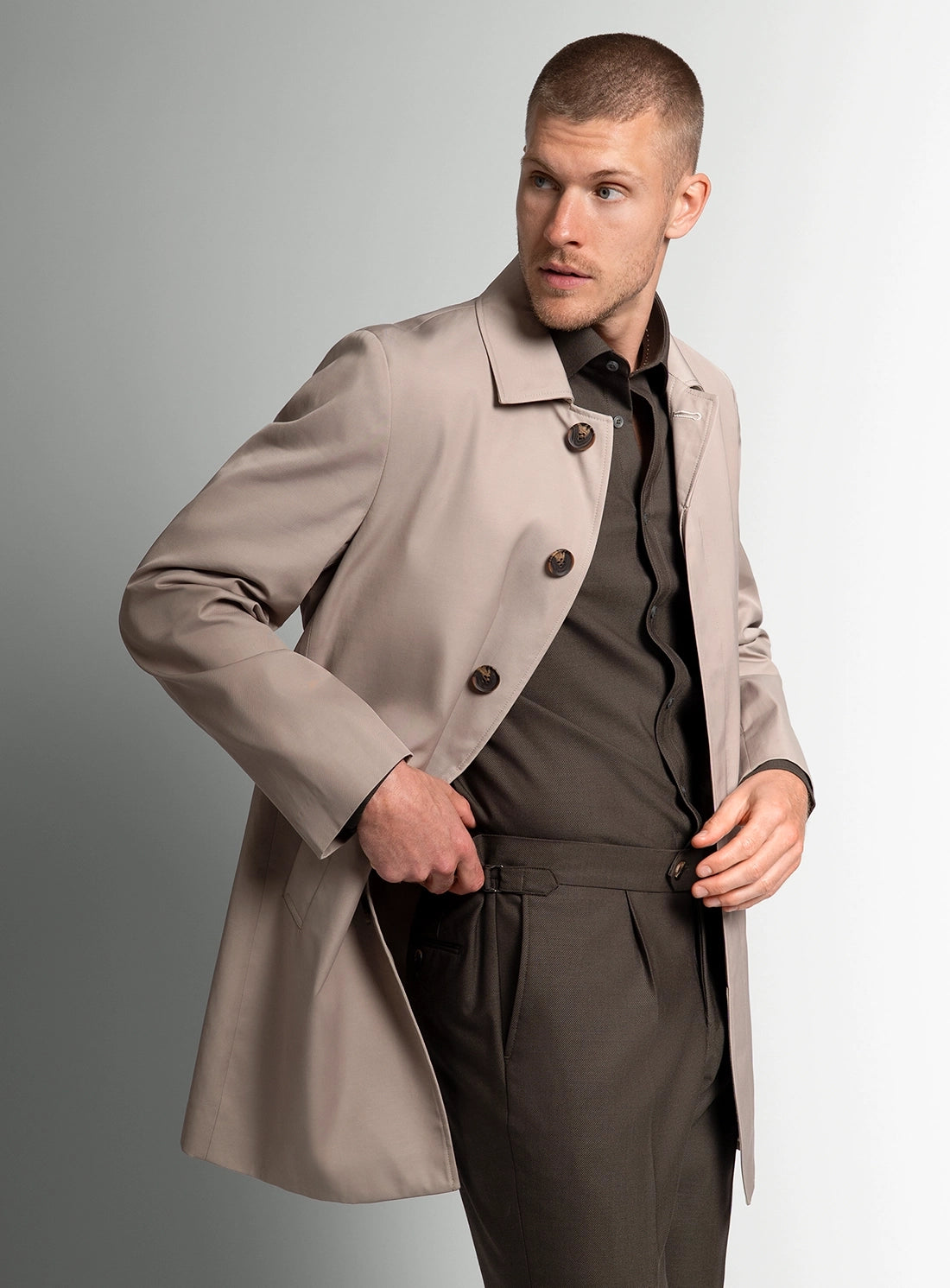 Man wearing a beige trench coat over a dark outfit on a gray background