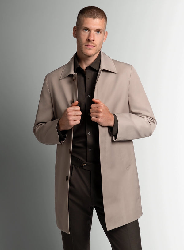 Man wearing a beige overcoat over a black shirt and pants on a gray background