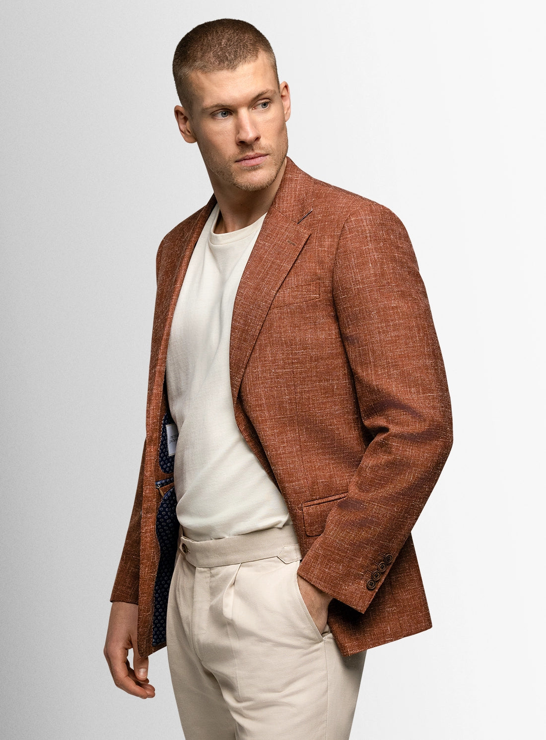 Man wearing a brown blazer over a white shirt and beige pants on a light gray background