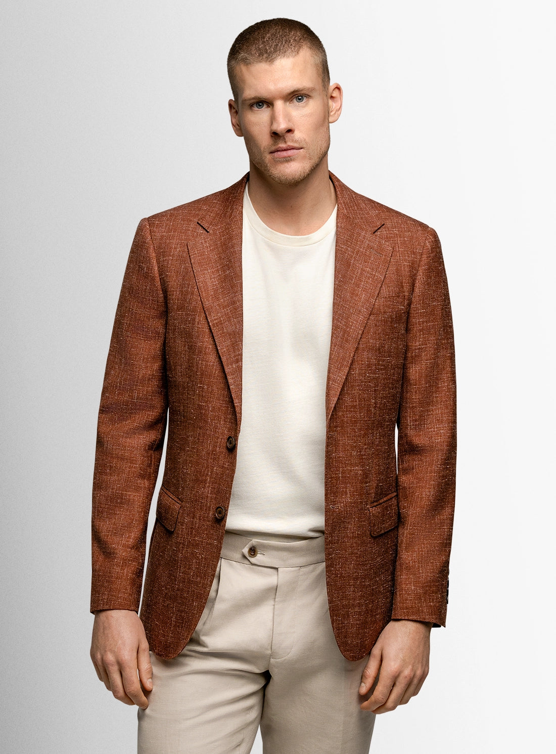 Man wearing a brown blazer over a white shirt and beige pants on a light gray background